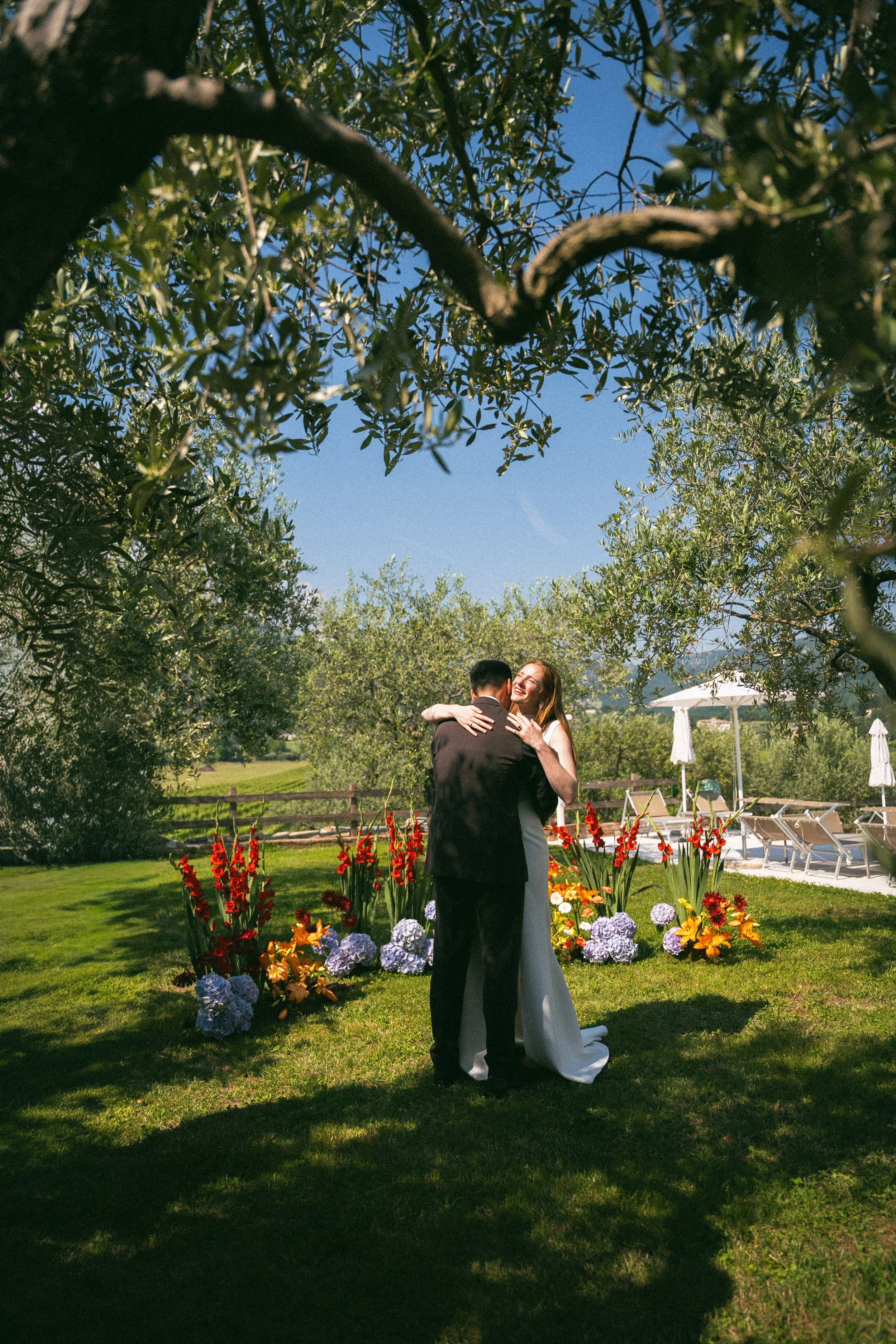 Una coppia che si sposa in un giardino con fiori colorati, sotto un albero e con un cielo sereno, mentre si abbracciano e sorridono.