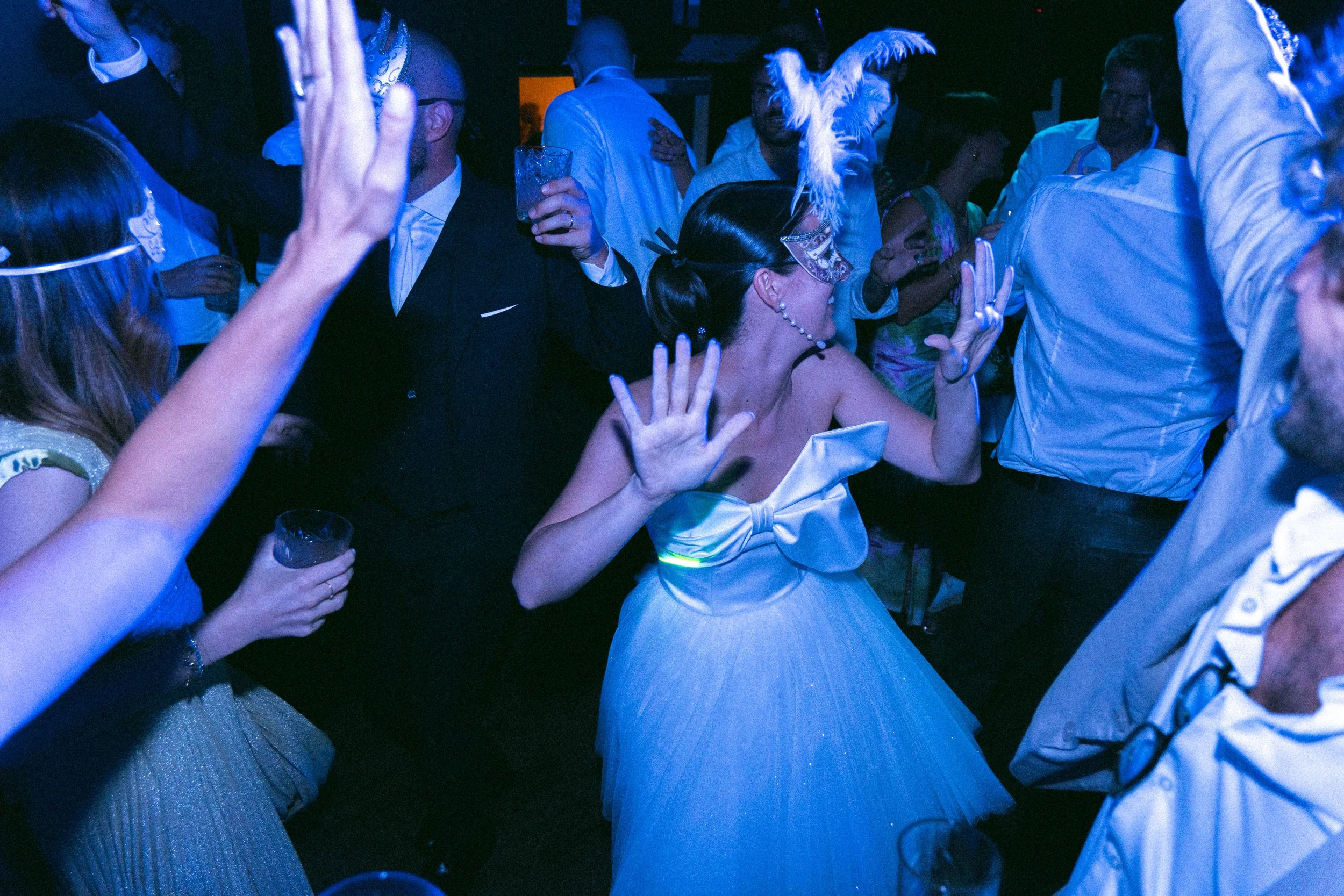 Persone che ballano e si divertono in un ambiente con illuminazione blu, alcune indossano maschere e accessori festivi.