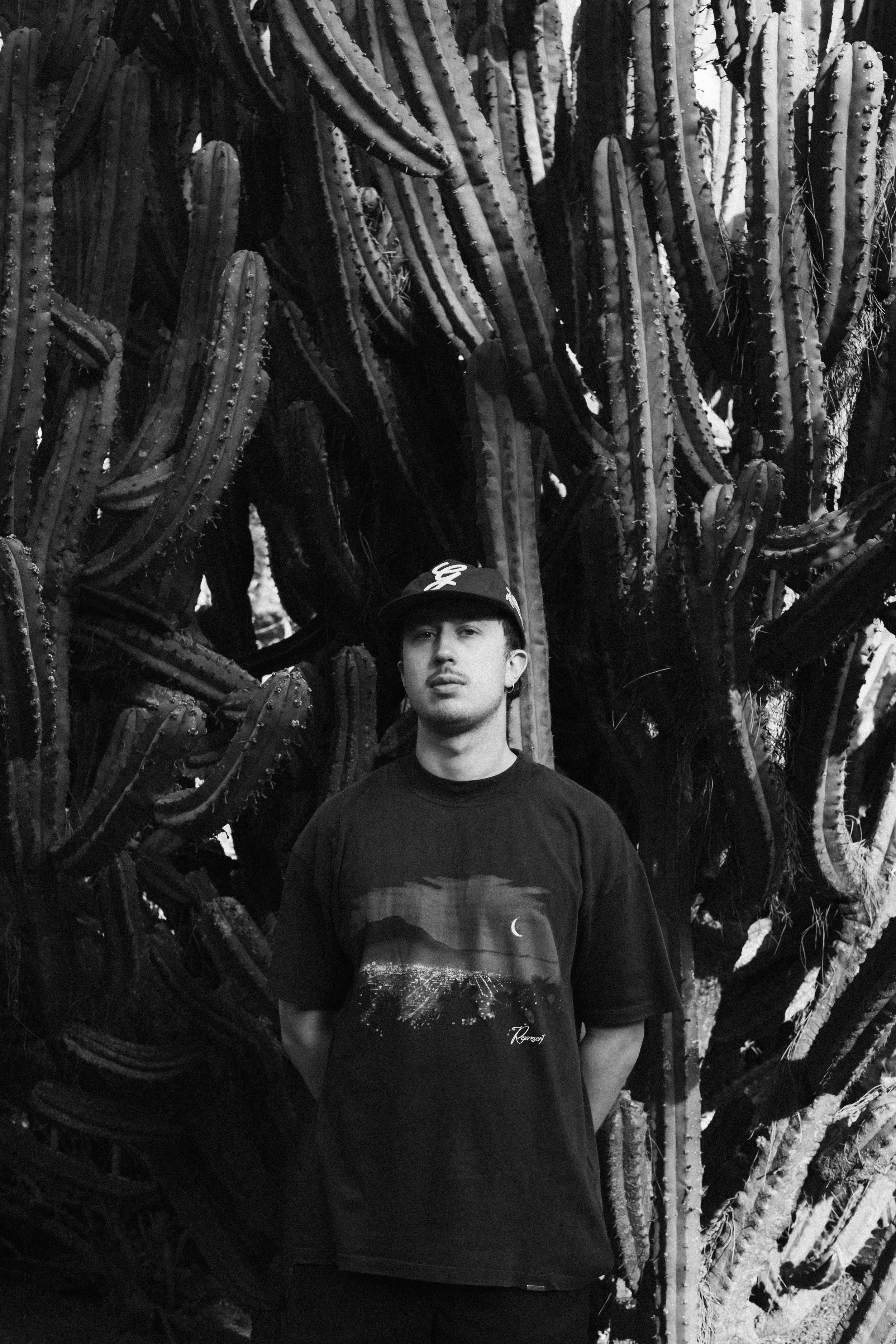 Un giovane uomo con cappellino nero e maglietta con un paesaggio di notte, tra cui un cielo stellato, un ponte e una luna, si trova in piedi davanti a un grande cactus.
