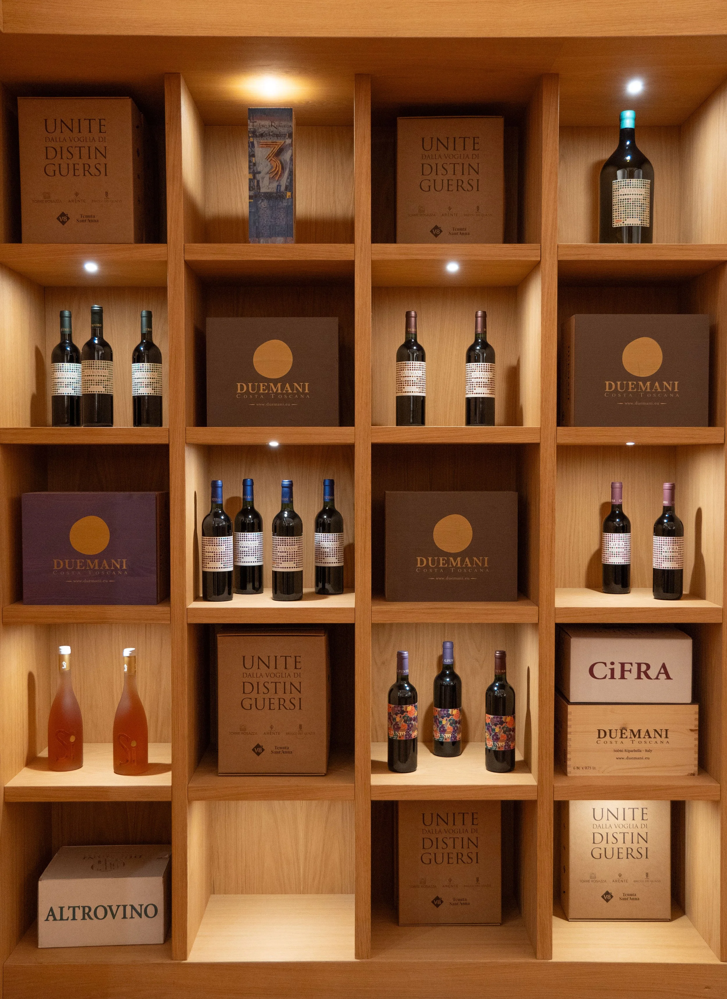 Scaffale con bottiglie di vino, scatole e decorazioni in legno, illuminazione a soffitto.