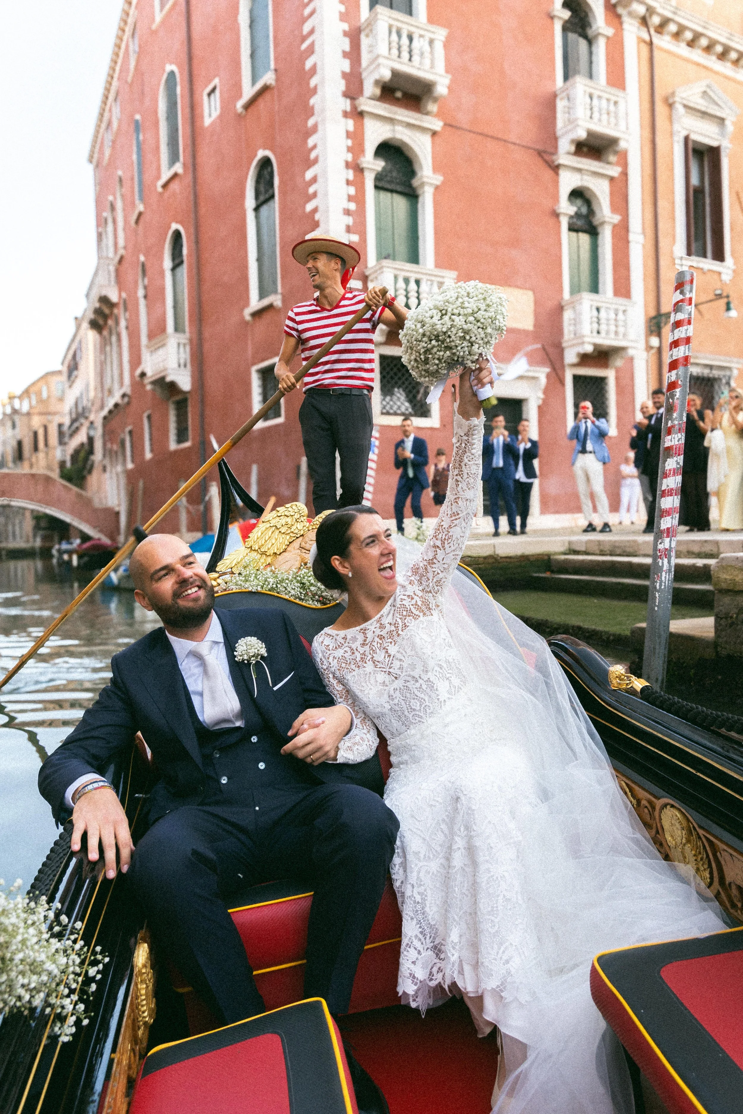 Coppia di sposi in barca a Venezia con il marito in abito formale e la moglie in abito da sposa, circondati da amici che festeggiano, sullo sfondo una palazzina veneziana rosa con dettagli architettonici.