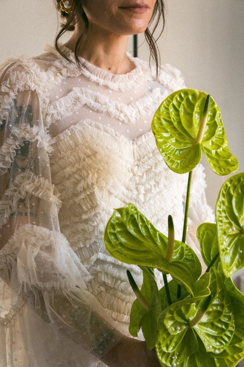 Donna con abito bianco ricamato e pizzo, che tiene una pianta verde con foglie grandi e lucide.
