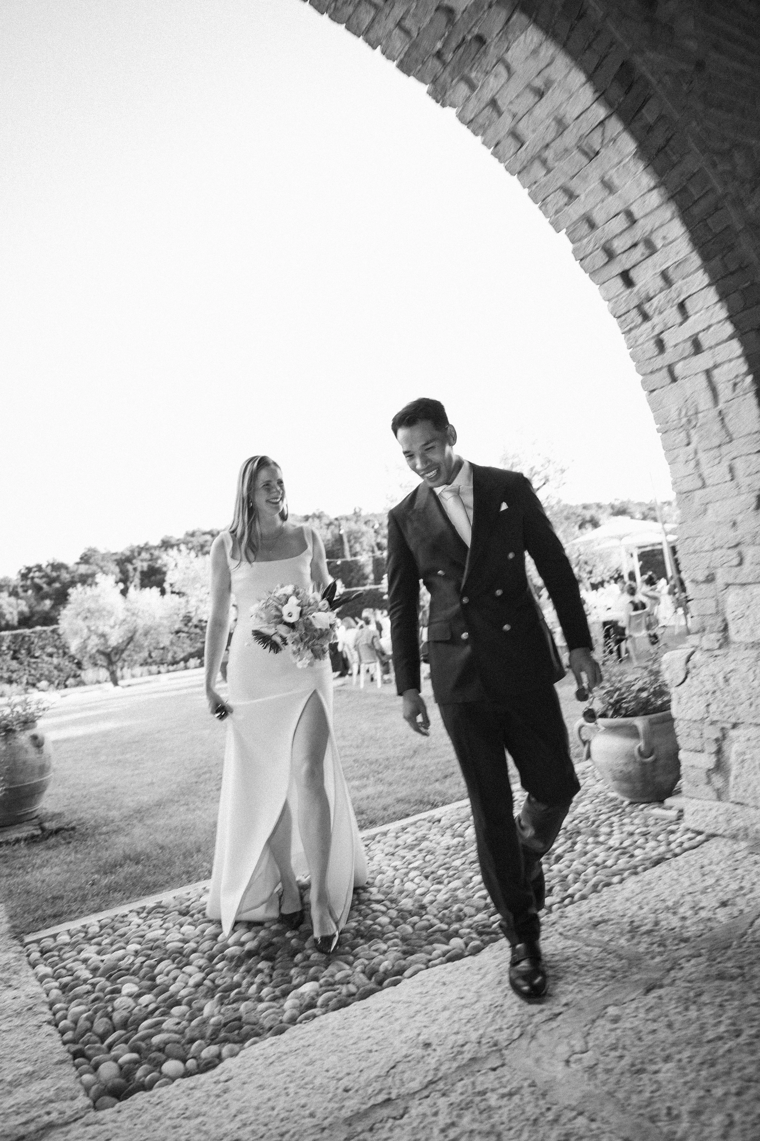Una coppia di sposi che cammina sorridendo in un giardino, con un arco di pietra sullo sfondo, in un giorno soleggiato.