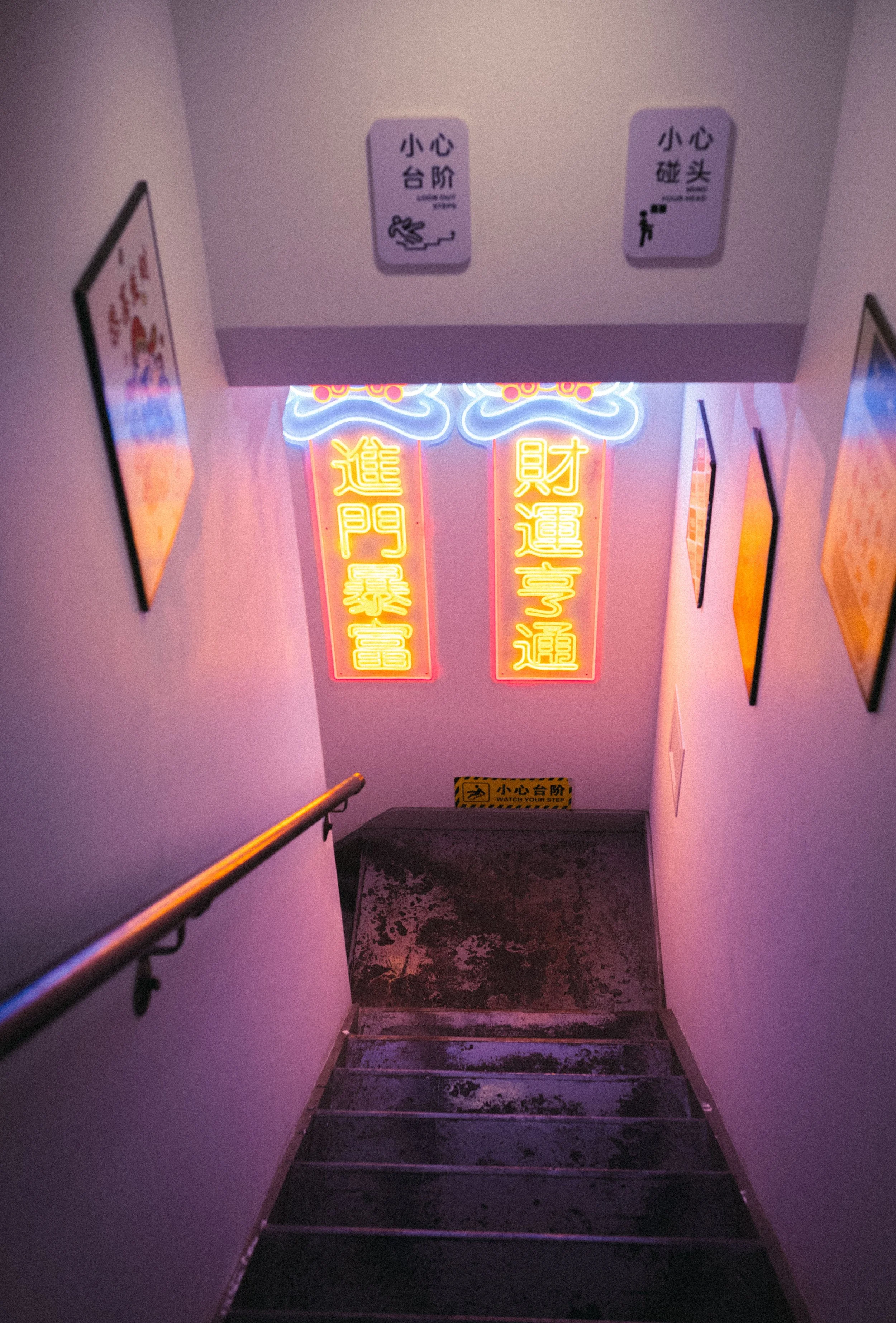 Scalinata in un edificio con segnali luminosi al neon gialli e rosa con scritte in cinese, cartelli di attenzione e segnali di sicurezza, pareti con poster.