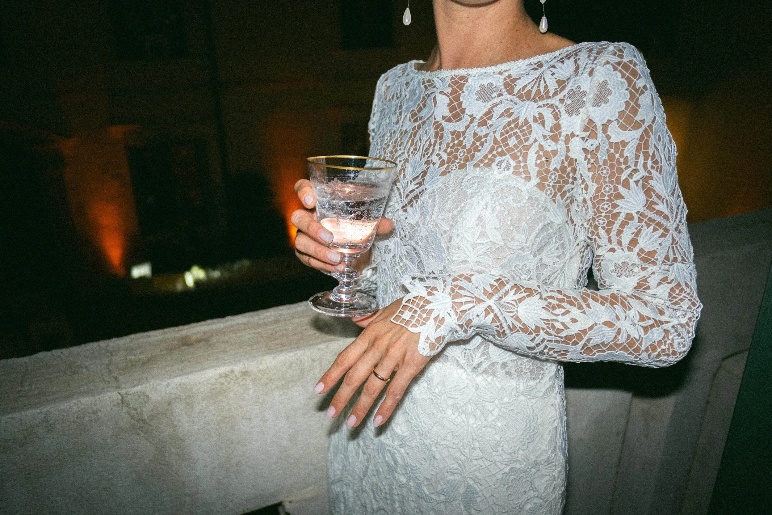 Donna con abito bianco in pizzo, portando un bicchiere di acqua, con orecchini pendenti e un anello sulla mano, sfondo scuro con luci calde.
