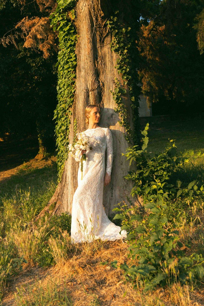 Una donna in abito da sposa bianco si appoggia a un grande albero in un'area verde, con foglie verdi e luce dorata del tramonto.