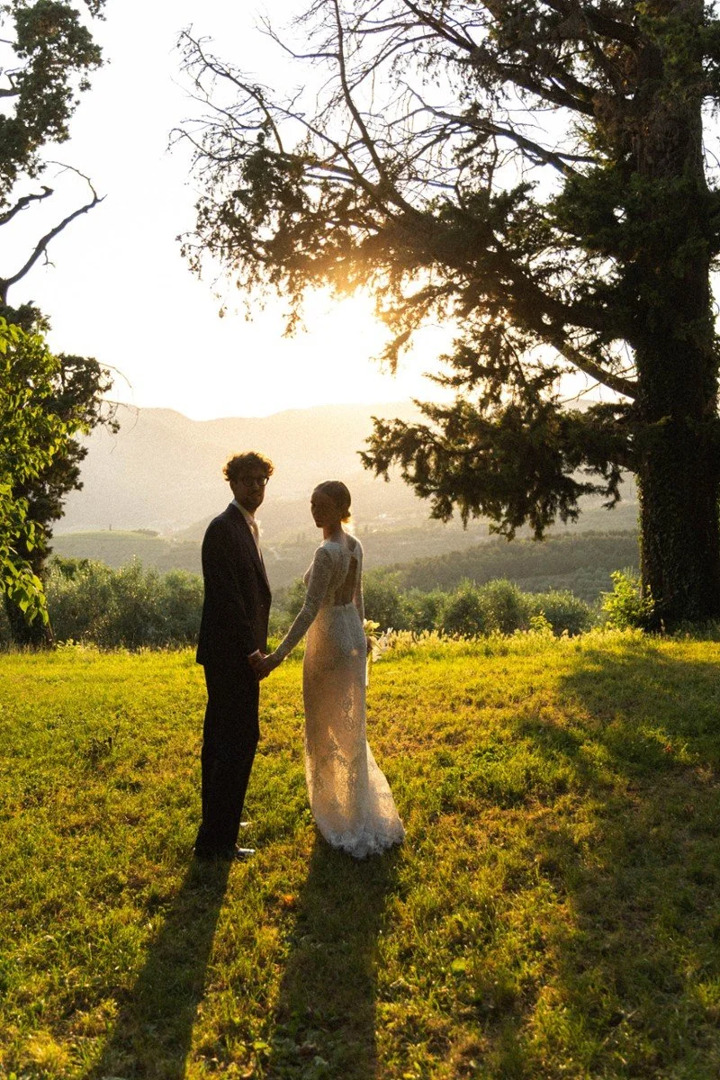 Coppia di sposi che si tiene per mano in un campo verde al tramonto, con alberi e montagne sullo sfondo.