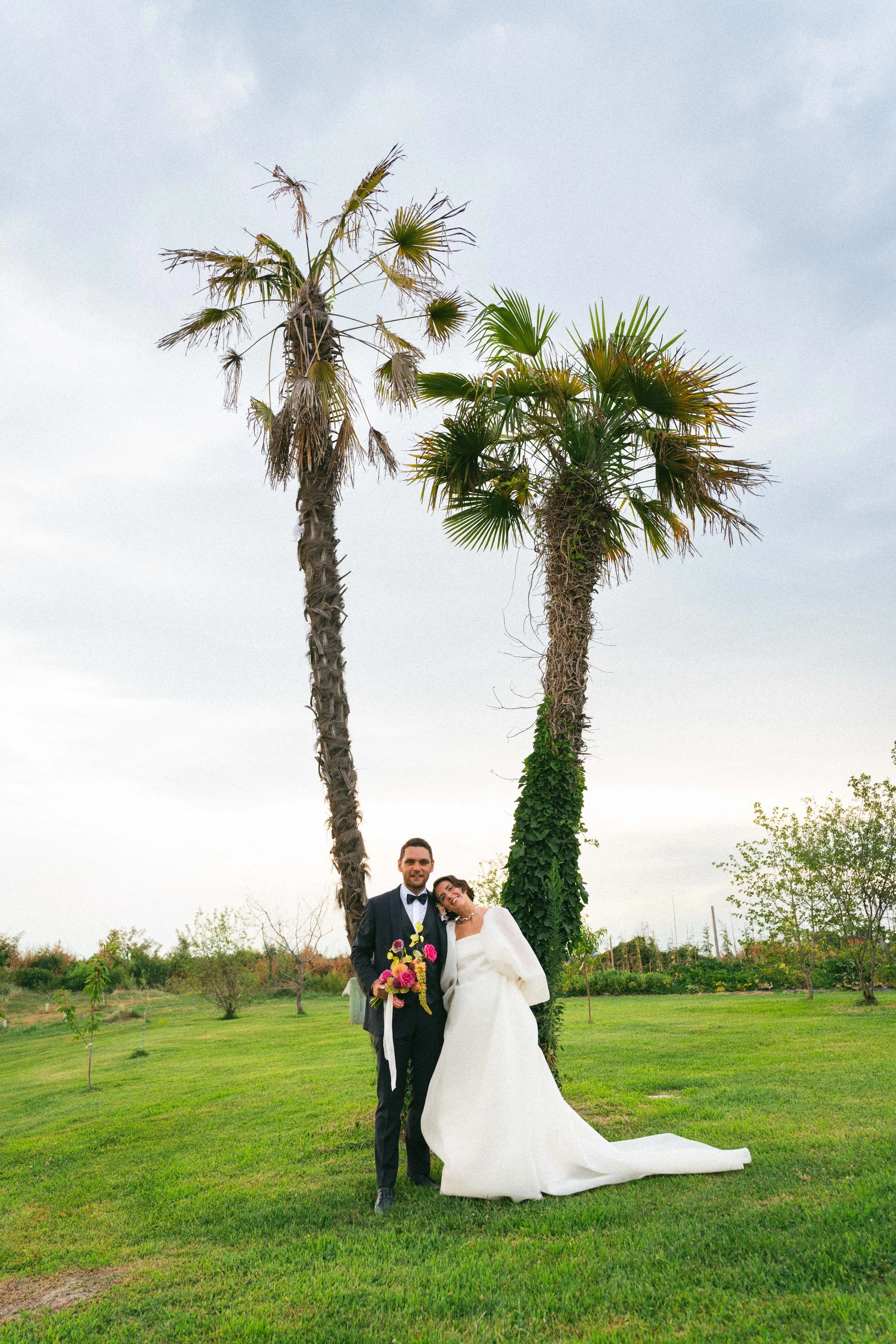 Una coppia di sposi in abiti da matrimonio, un uomo in completo nero con papillon e donna in abito bianco lungo, sorridenti, in piedi in un campo verde con due palme miste, una secca e una più verde e rigogliosa.