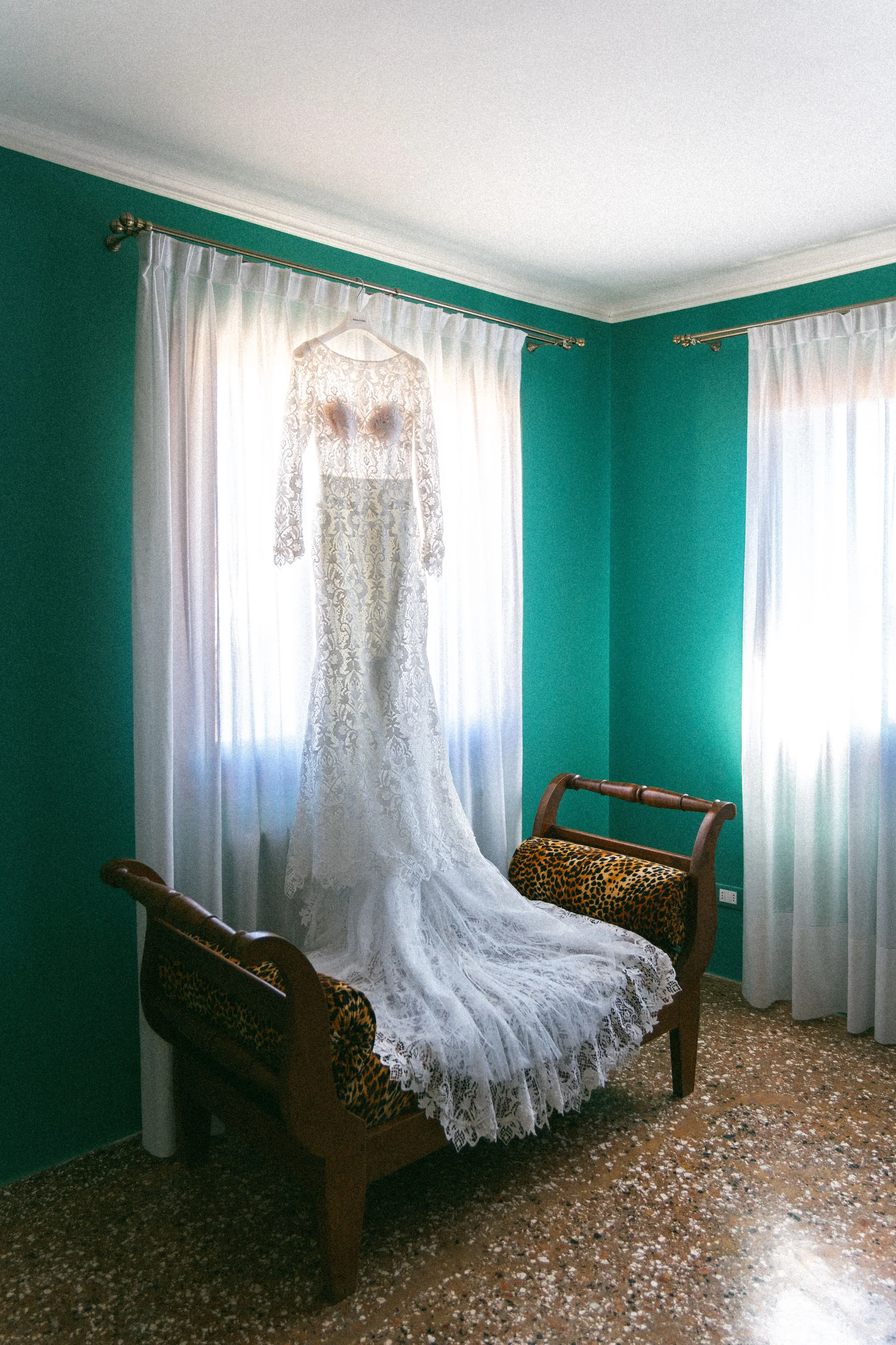 Una stanza con pareti verde smeraldo, due tende di tessuto leggero e una gonna da sposa di pizzo appesa alla finestra. Una panca con rivestimento leopardato si trova sotto la finestra, con una struttura di legno scuro.