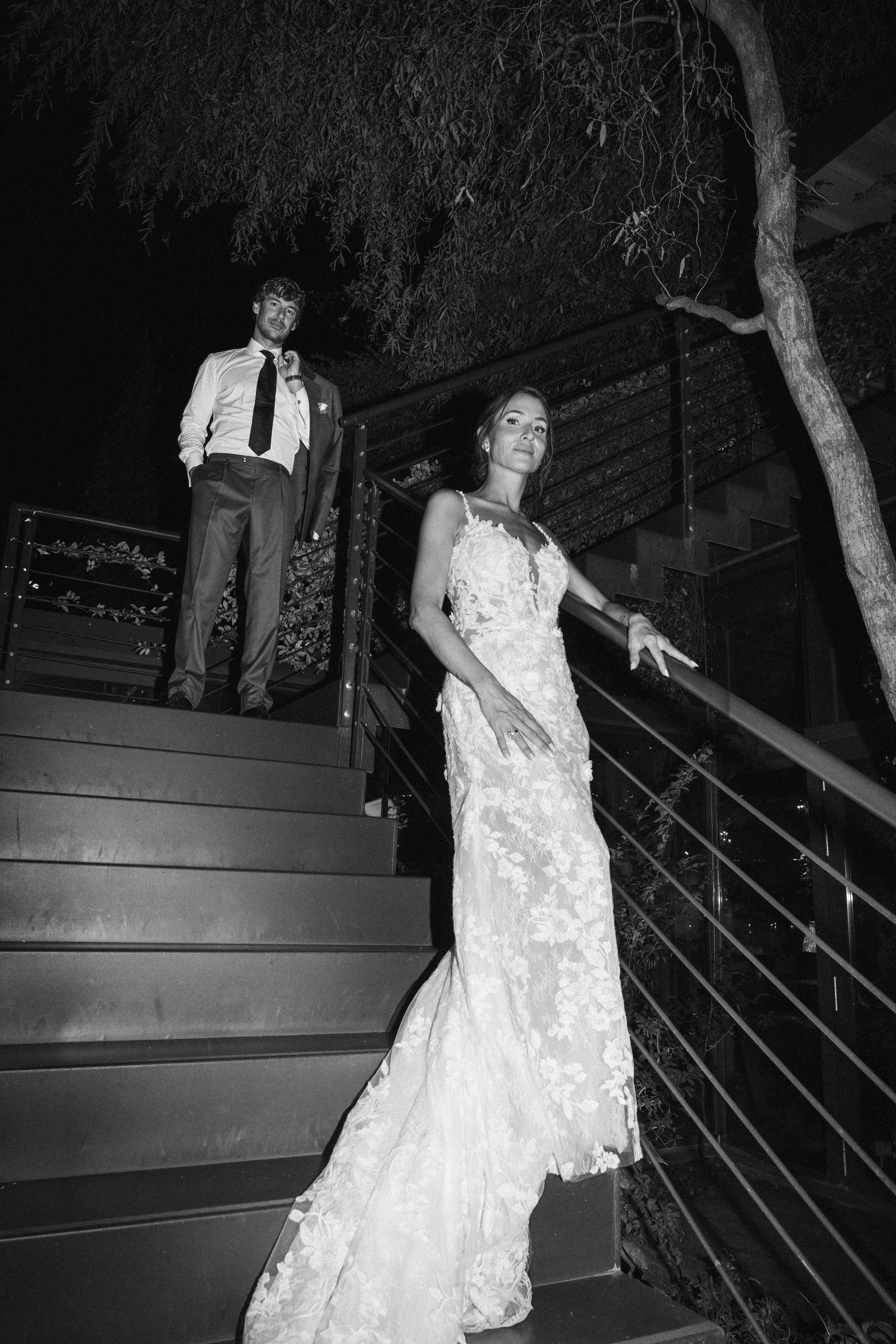 Una coppia di sposi in abiti da nozze, una donna in un vestito bianco lungo e un uomo in abito scuro con camicia bianca e cravatta, posano su uno scalinate di notte con alberi intorno.