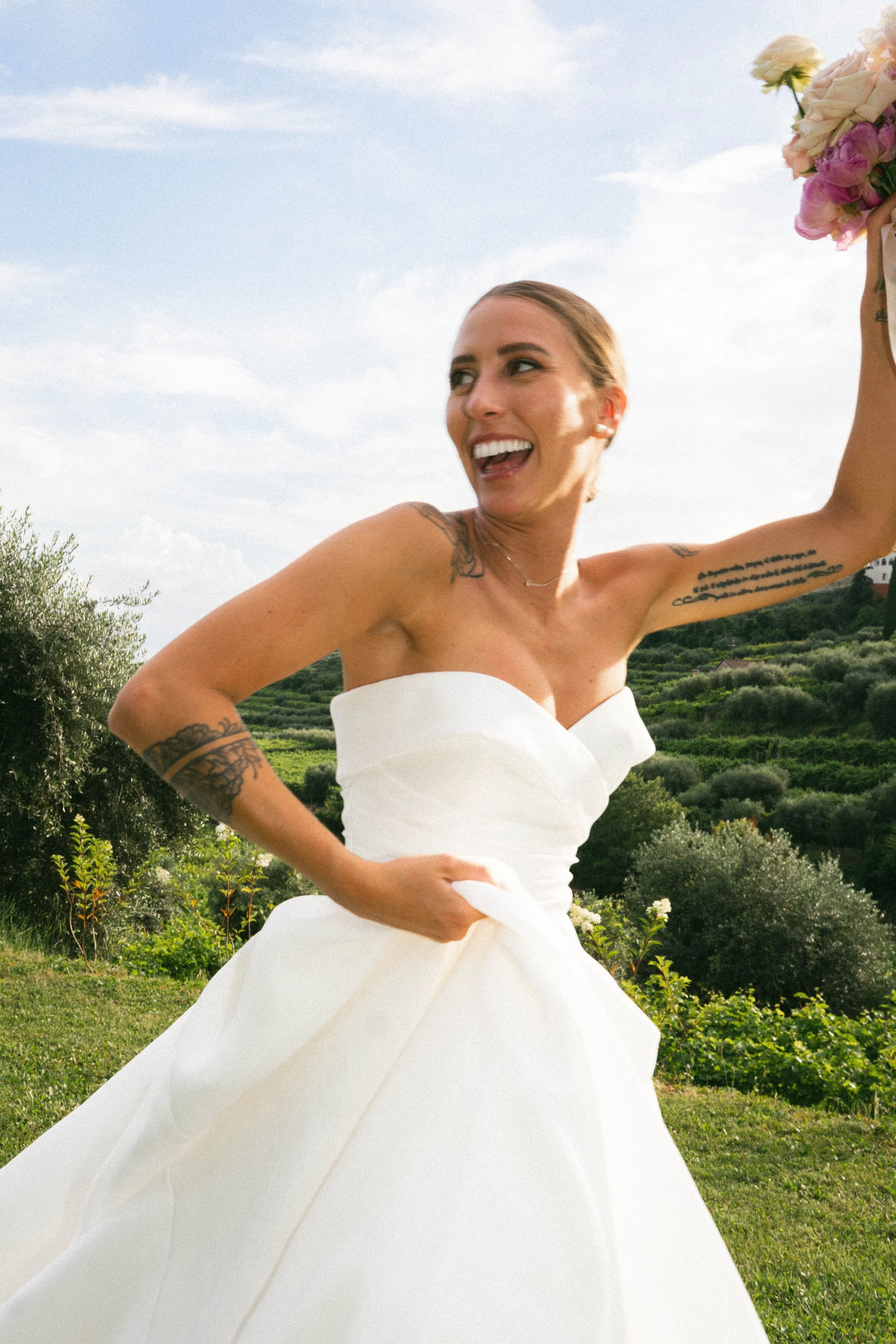 Una donna sorridente in abito bianco in un campo verde con colline e cielo nuvoloso sullo sfondo, che tiene un bouquet di fiori.