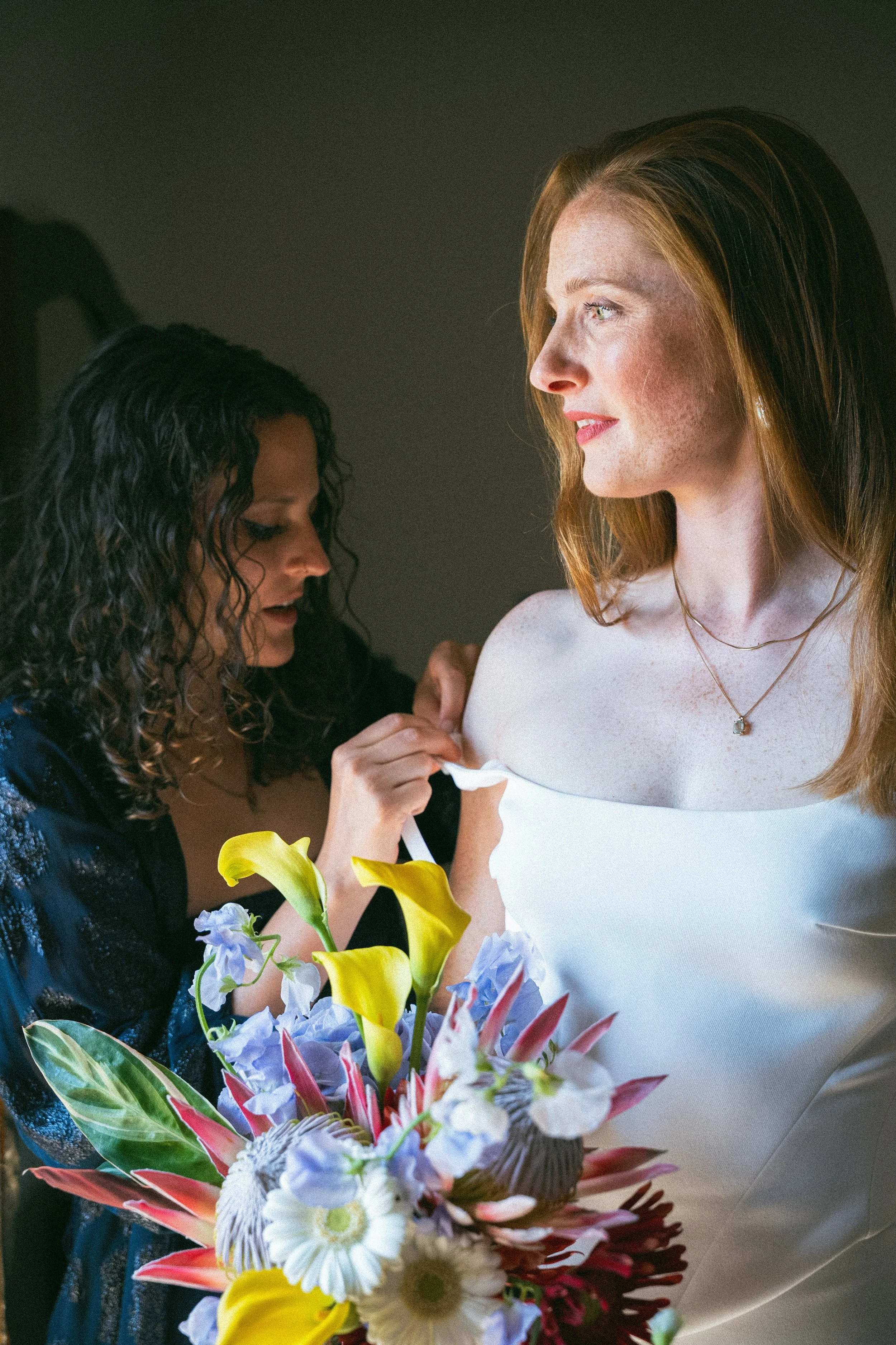 Una donna con capelli castani che indossa un abito bianco e una collana, riceve un bouquet di fiori colorati da un'altra donna con capelli ricci scuri, sullo sfondo scuro.
