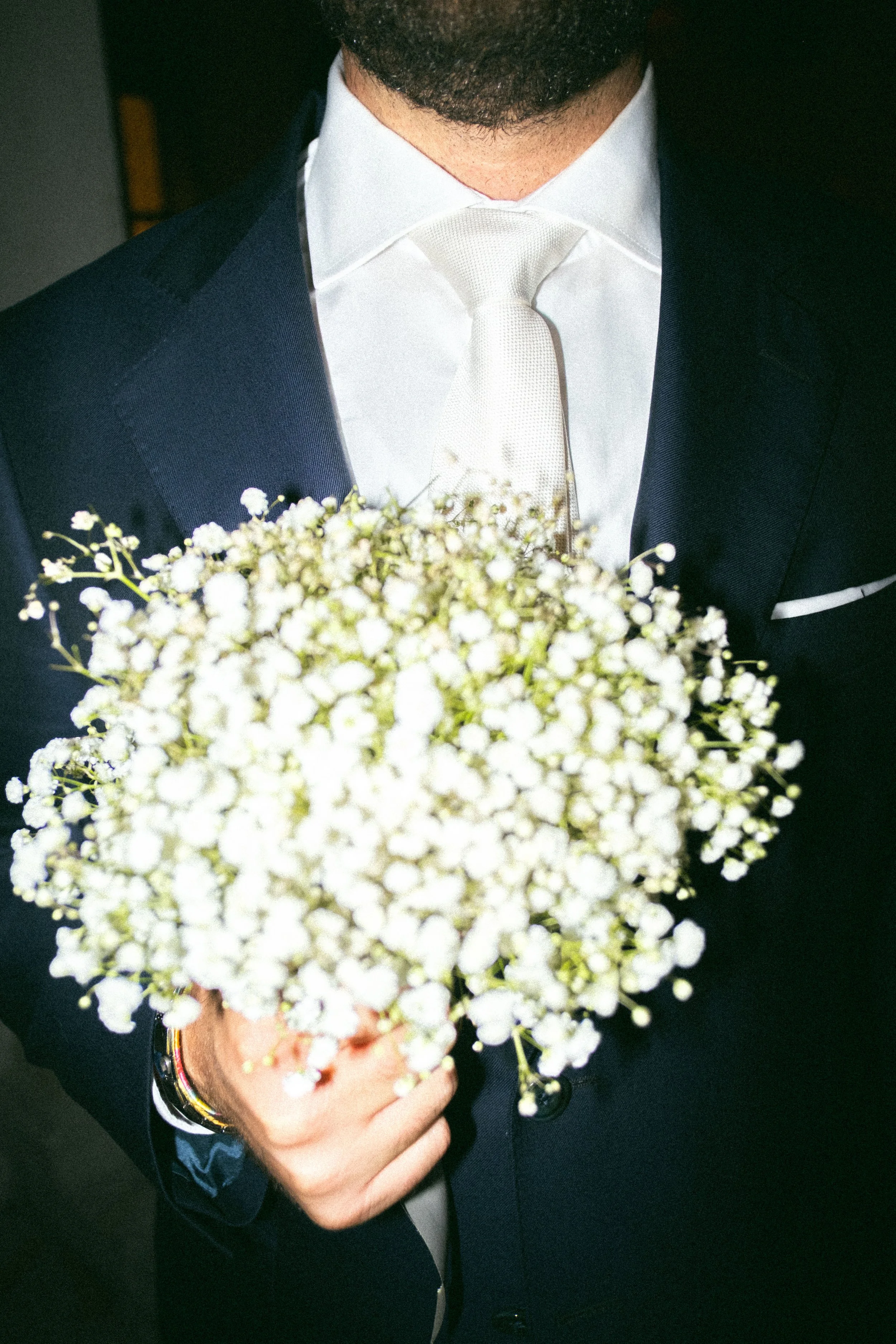 Uomo vestito elegantemente con giacca e cravatta, che tiene un mazzo di fiori bianchi davanti al volto.