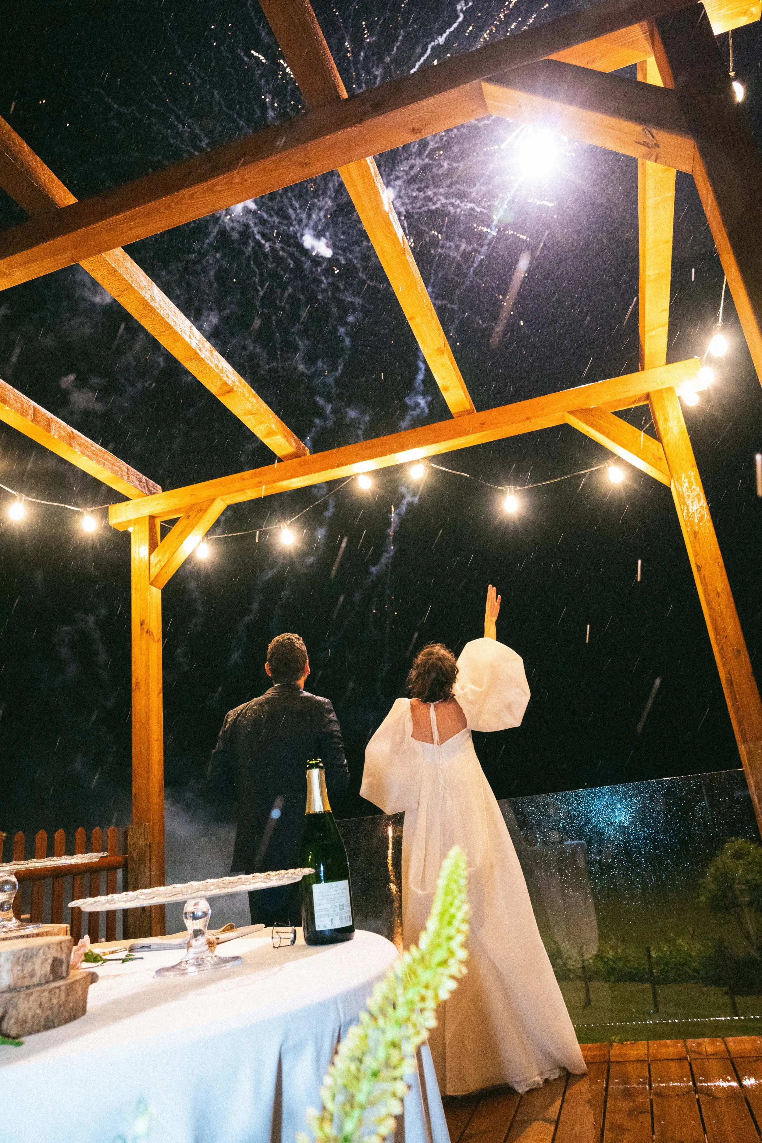 Una cerimonia di matrimonio all'aperto sotto una tettoia di legno, con il cielo stellato visibile sopra. Gli sposi sono rivolti lontano, uno indossa un abito bianco e l'altro un abito scuro. Sul tavolo di prima piano ci sono bottiglie di vino e ogget