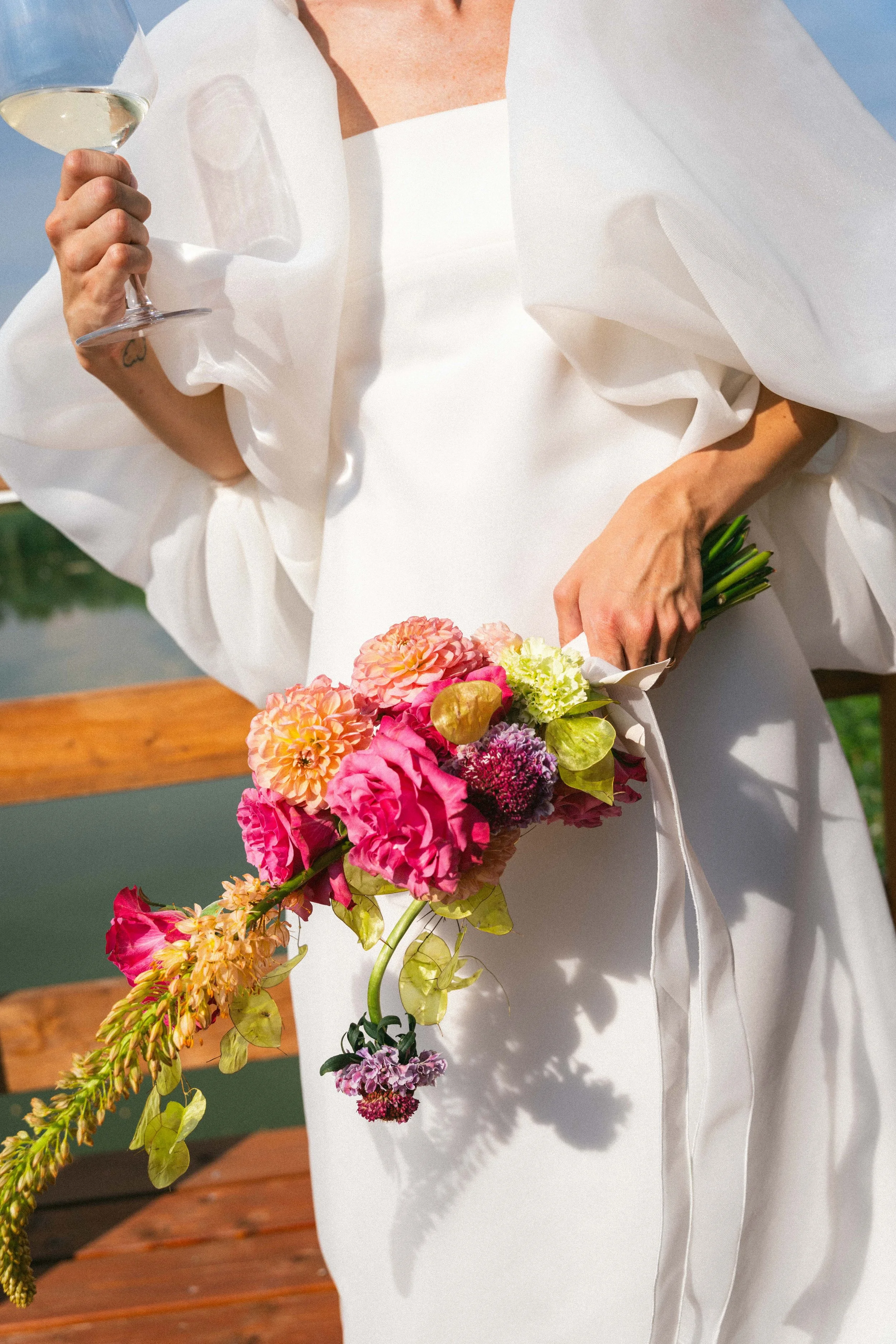 Donna in abito bianco con bouquet di fiori colorati e bicchiere di vino bianco in mano