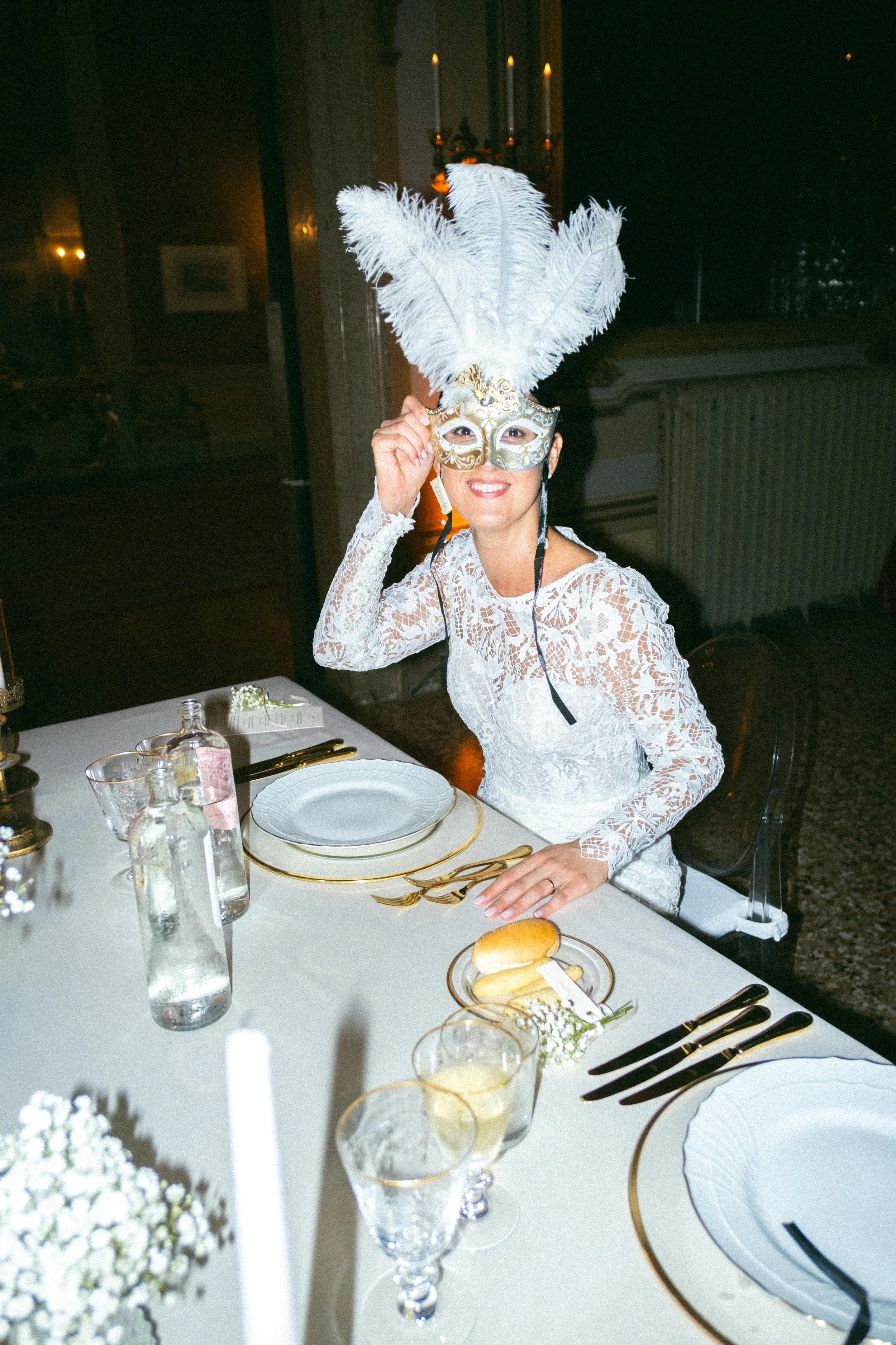 Una donna con un costume da carnevale, indossa una maschera decorata e un grande copricapo di piume bianche, seduta a tavola apparecchiata per una cena elegante.