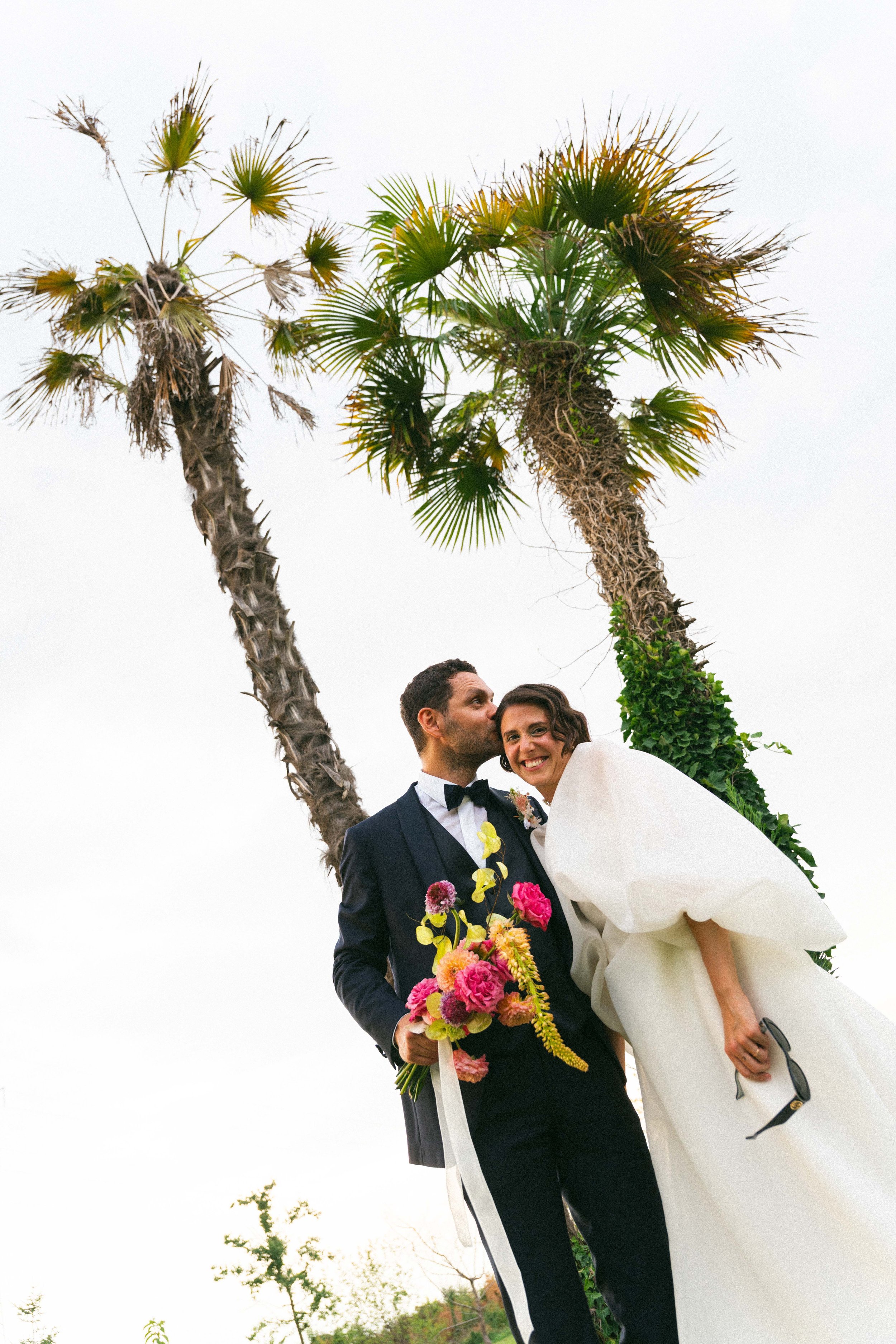 Una coppia di sposi abbracciati davanti a due palme. L'uomo indossa un abito scuro con papillon, la donna un vestito bianco con fiore all'occhiello. Sorridono, con un bouquet di fiori colorati in mano.
