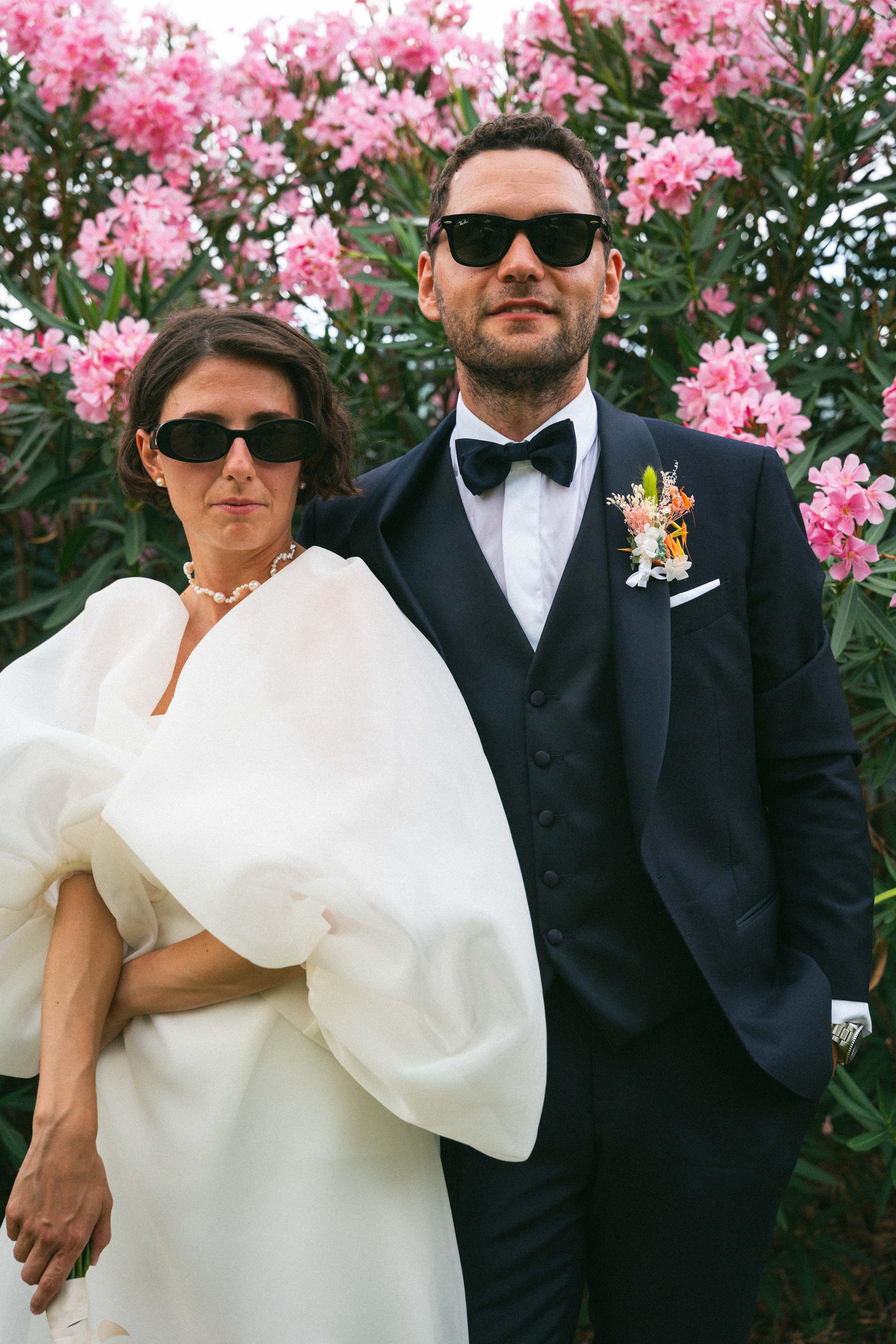 Una coppia elegante, un uomo e una donna, indossano occhiali scuri e sono vestiti in abiti da cerimonia. La donna porta un abito bianco con dettagli voluminosi e una collana di perle, mentre l'uomo indossa uno smoking nero con papillon e una boutonni