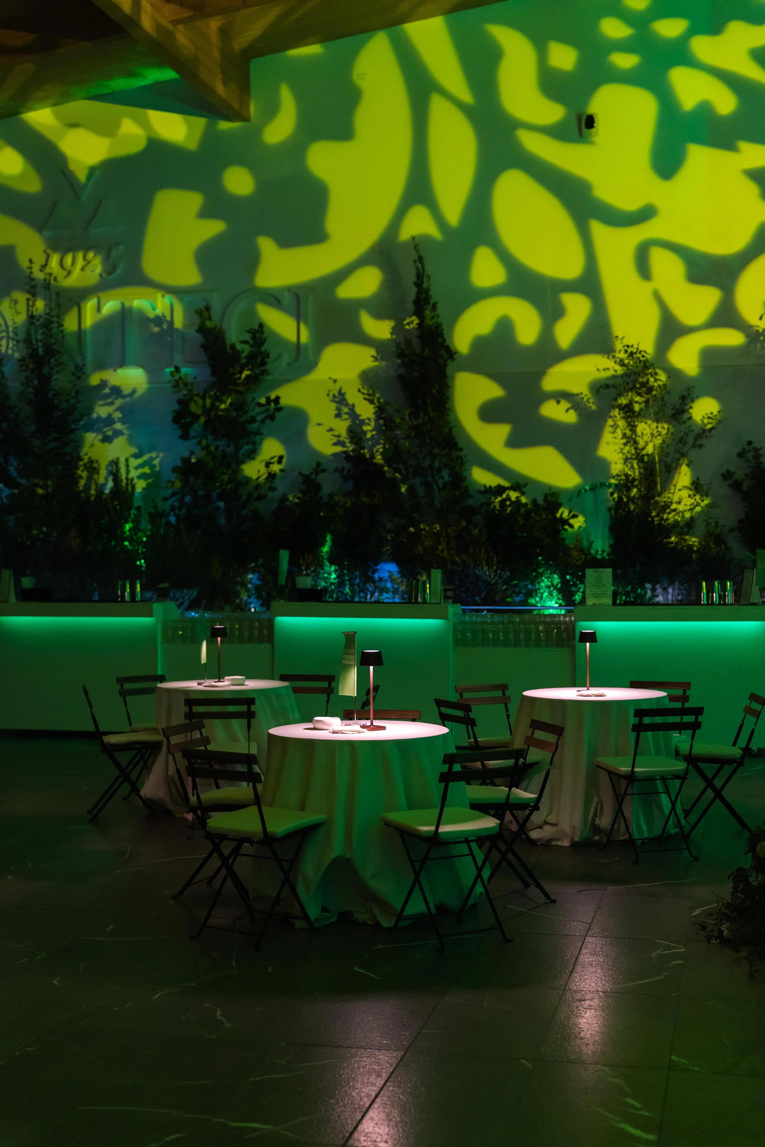Sala con tavoli rotondi con tovaglie bianche, sedie nere con cuscini chiari, illuminazione soffusa con luci da tavolo, parete con proiezioni di luci verdi e gialle, piante sullo sfondo.