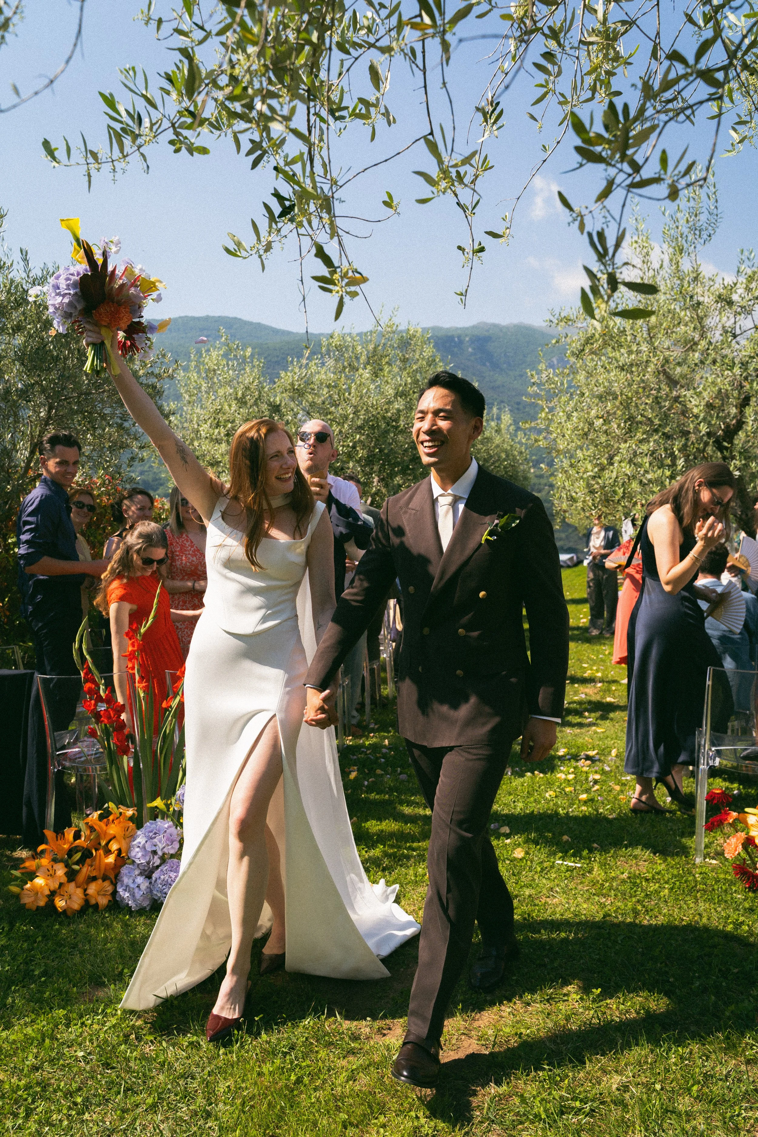 Una coppia di sposi felice che si tiene per mano durante il matrimonio all'aperto, con molti invitati intorno e un paesaggio naturale con montagne sullo sfondo.