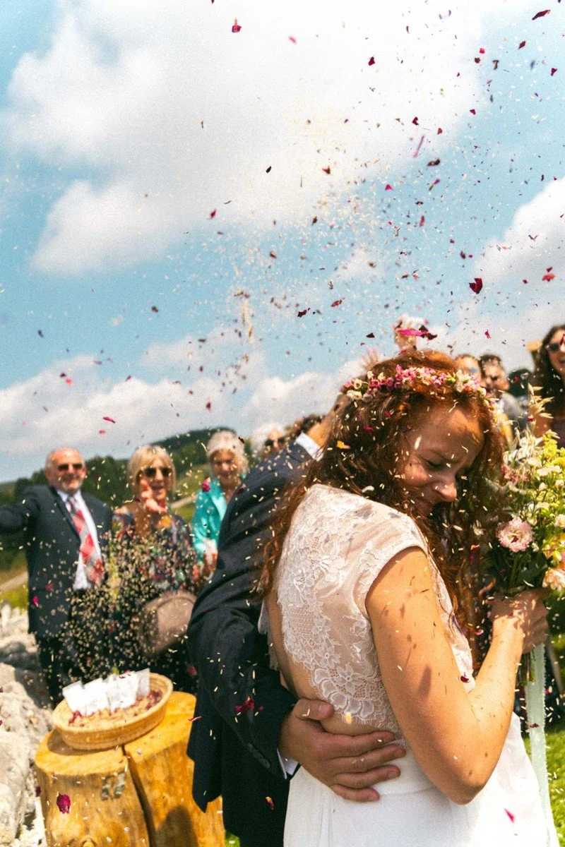 Una coppia di fidanzati, probabilmente in un matrimonio, che si scambia un bacio sotto una pioggia di petali di fiori, con un gruppo di persone sorridenti sullo sfondo e un cielo nuvoloso.
