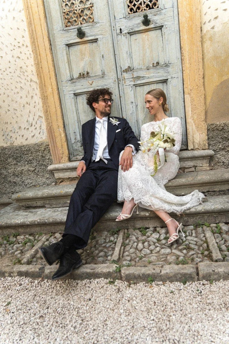Una coppia di sposi seduti su dei gradini di pietra davanti a una porta in legno bianco, sorridenti e in atteggiamento affettuoso.