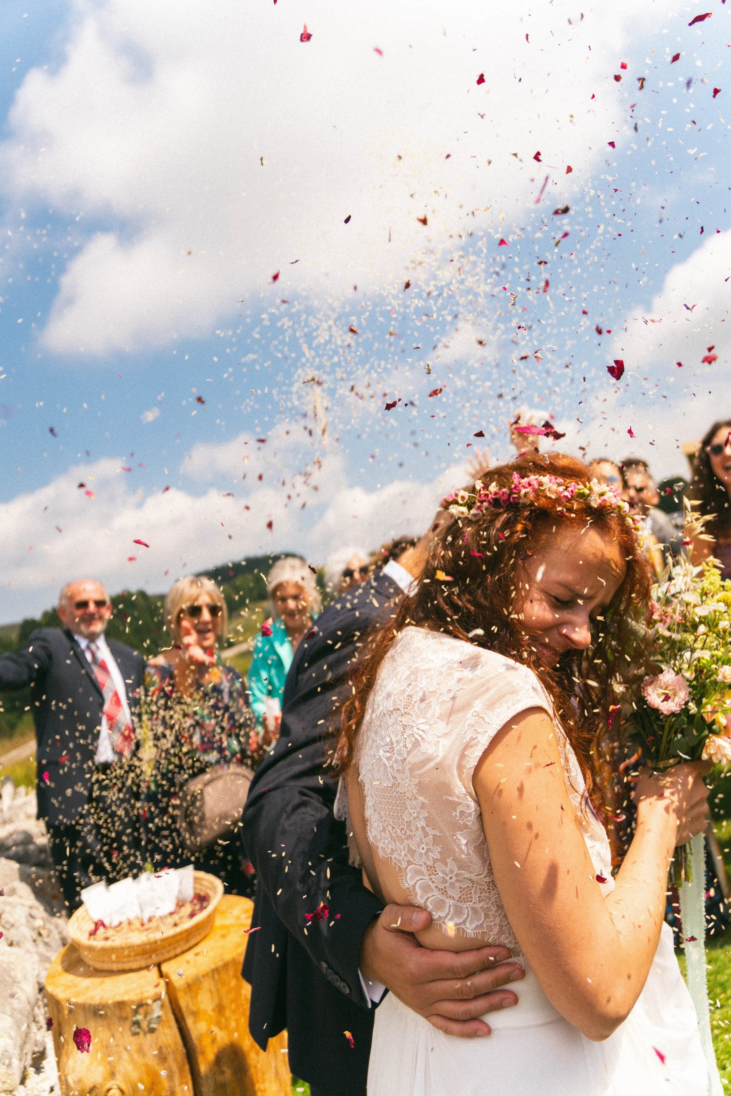 Una coppia di sposi si abbraccia durante una cerimonia all'aperto con bandiere di fiori e confetti nel cielo, celebrando il loro matrimonio.