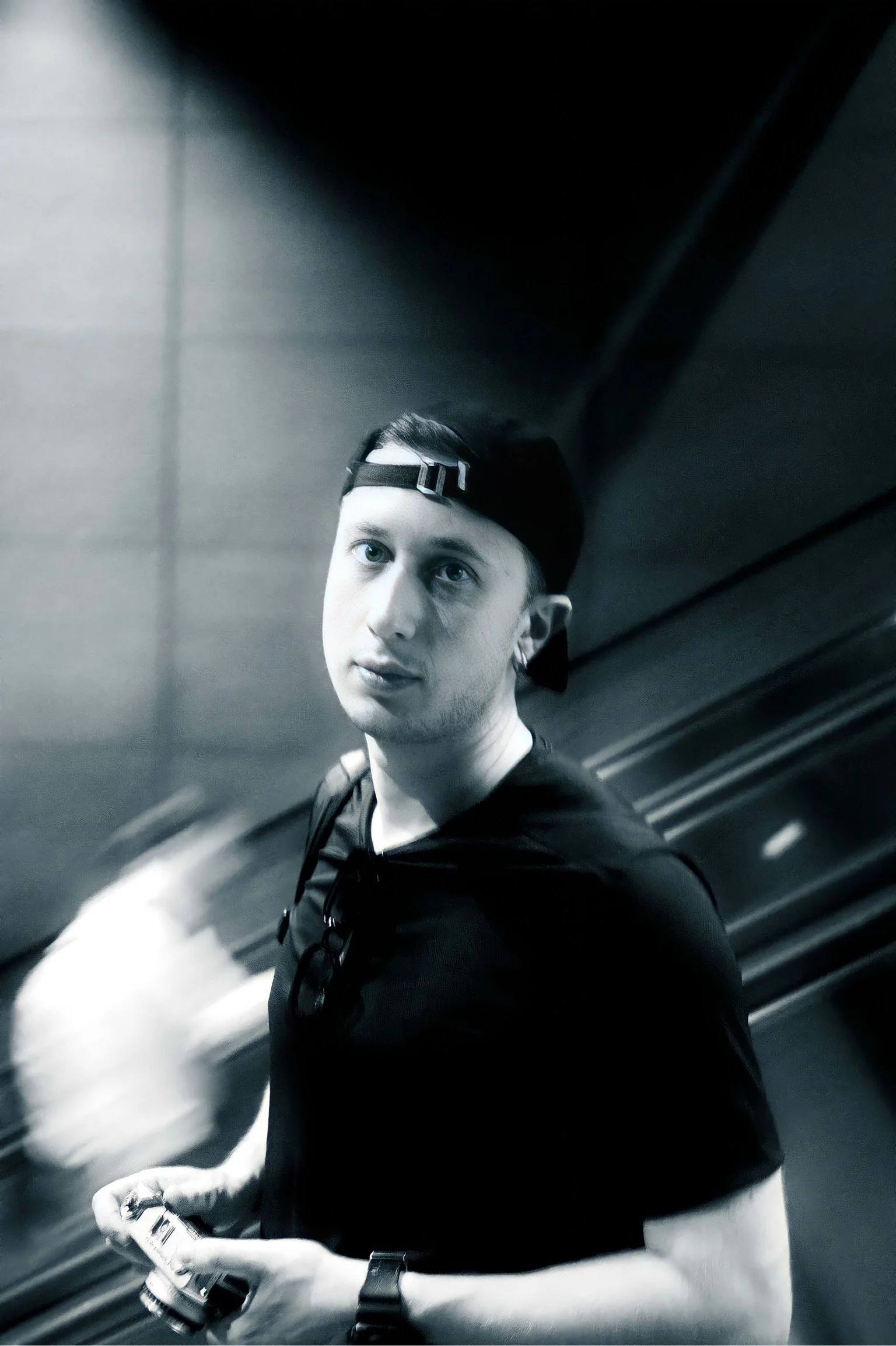 Ragazzo con cappellino rivolto all'indietro, che guarda verso l'obiettivo, tiene un telefono in mano e indossa una maglietta nera, in un ambiente con pareti di metallo