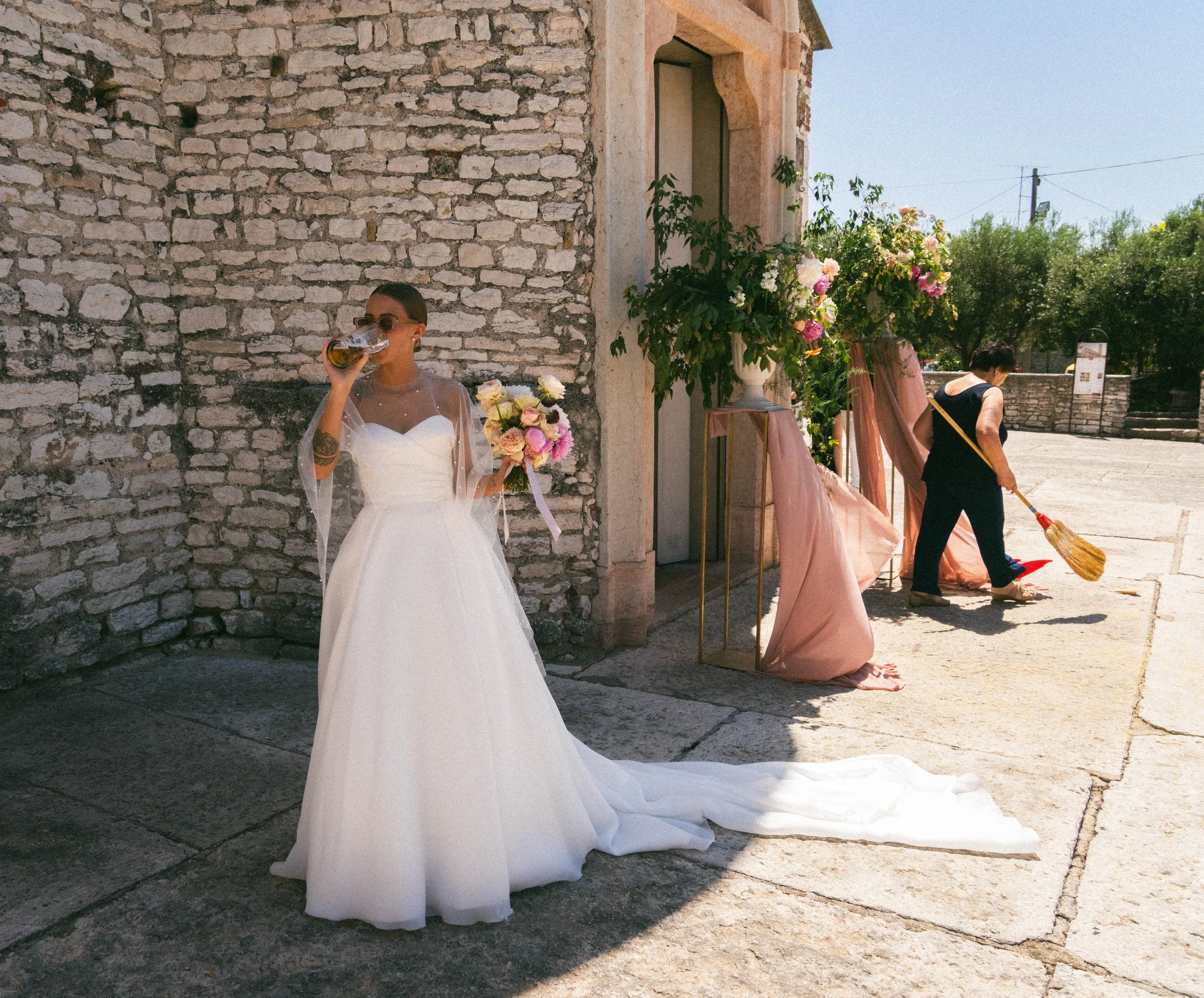 Donna in abito bianco con bouquet di fiori mentre beve una birra in un ristorante all'aperto, con muro di pietra e fiori sullo sfondo.