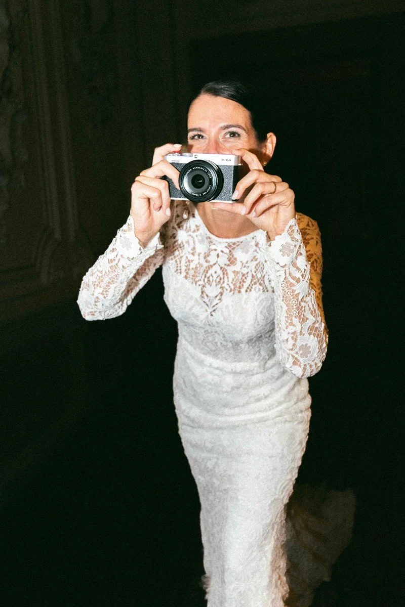 Una donna in abito bianco con ricami prende una foto con una macchina fotografica digitali, sorridendo.