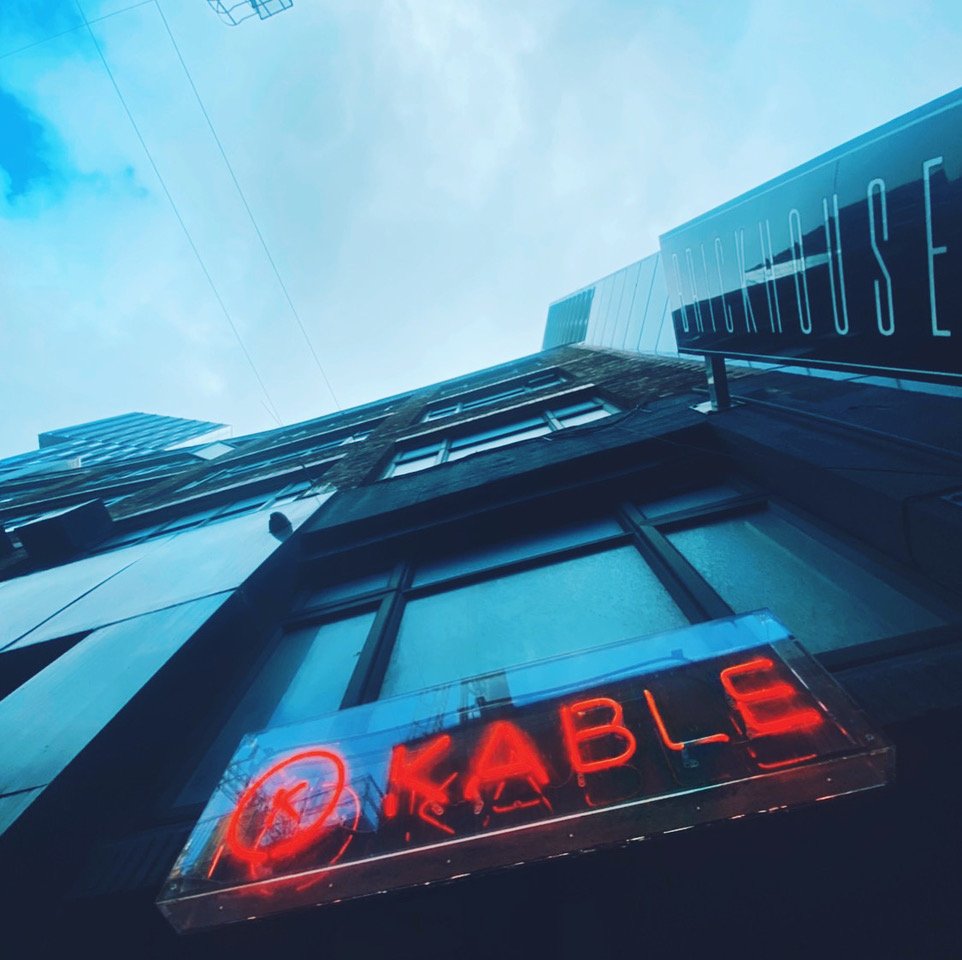 View of a tall building from below with neon signs that read "KABlE" and partly visible sign on the side of the building. Overcast sky overhead.