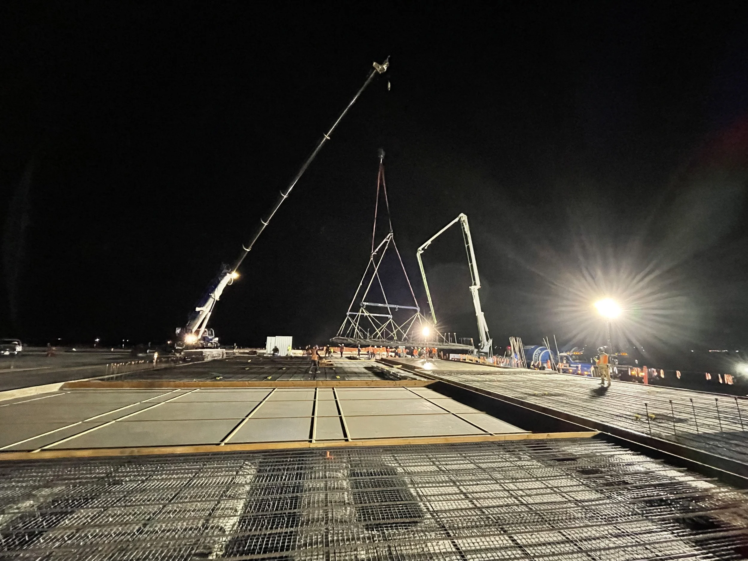 Nighttime construction scene on a bridge with workers, cranes, and equipment installing a large steel structure
