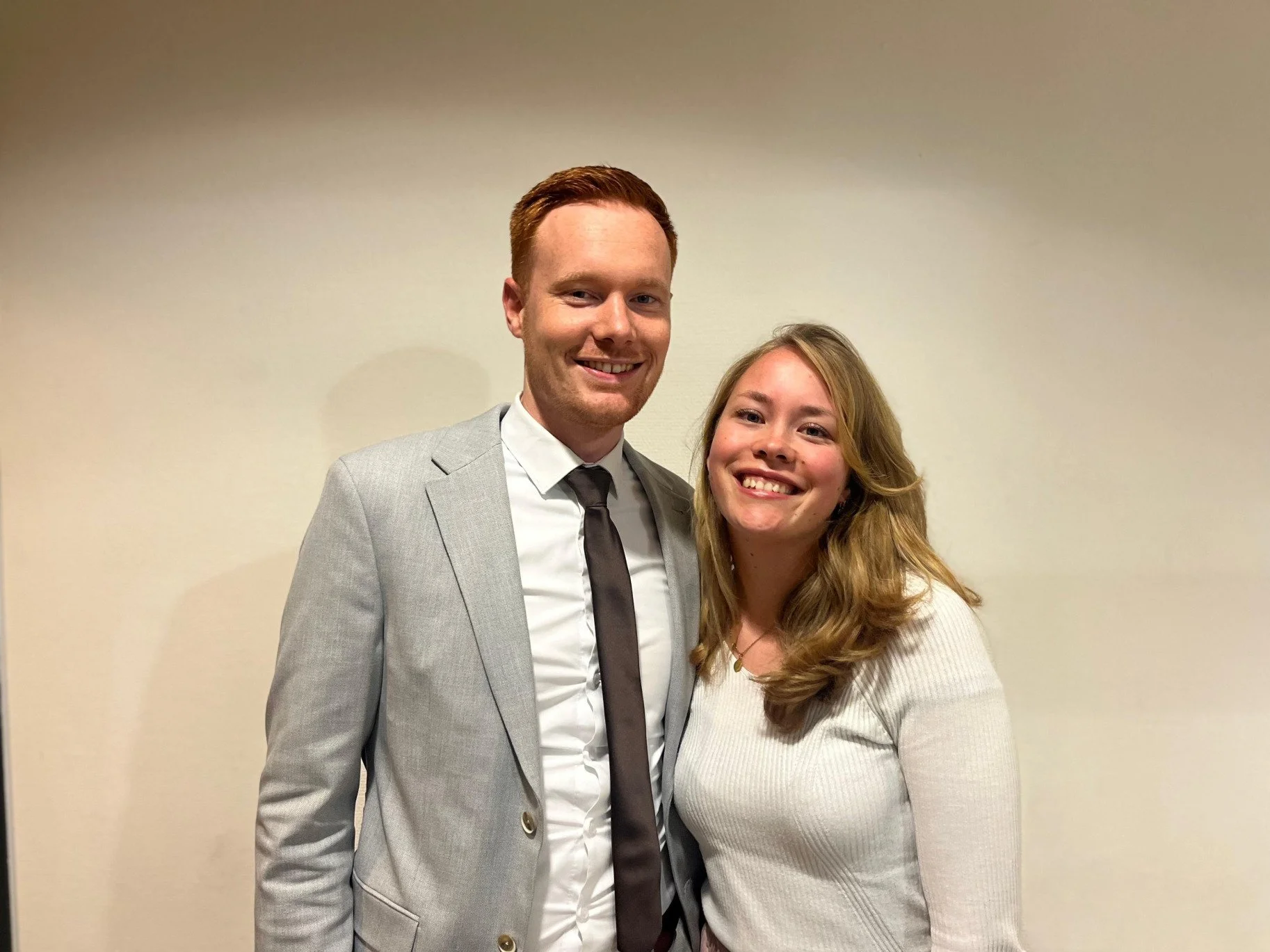 Een man in een grijs pak met witte blouse en zwarte stropdas staat naast een lachende vrouw met lang, golvend blond haar, beiden voor een effen lichtkleurige achtergrond.
