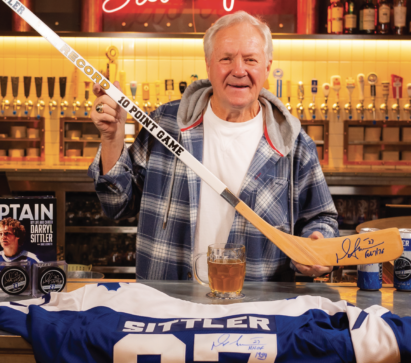 Darry Sittler now days holding a hockey stick with a signature, in a bar with beer taps in the background. The table in front has a signed hockey jersey, a mug of beer, and hockey pucks.