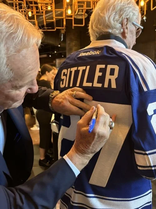The Hockey Legend Darryl Sittler signs a football jersey for another person. The jersey has 'SITTLER' written on the back and a large number 7. The setting is indoors with warm lighting and other people in the background.