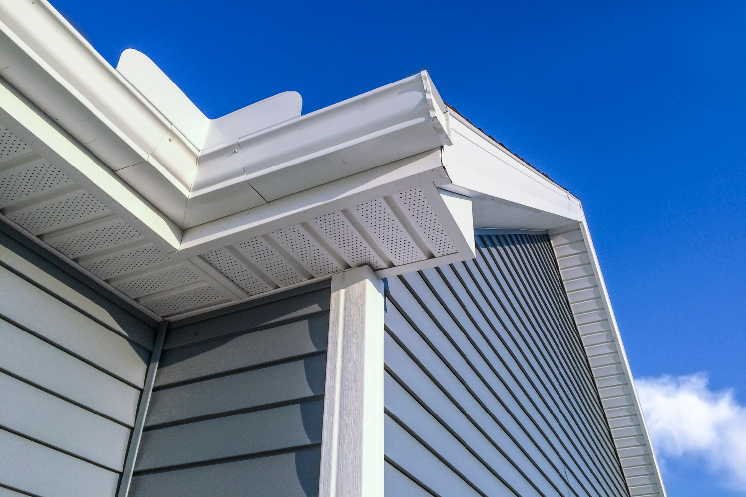Close-up of the corner of a house with gray siding and white trim, showing the eaves and gutter system against a blue sky with some clouds.