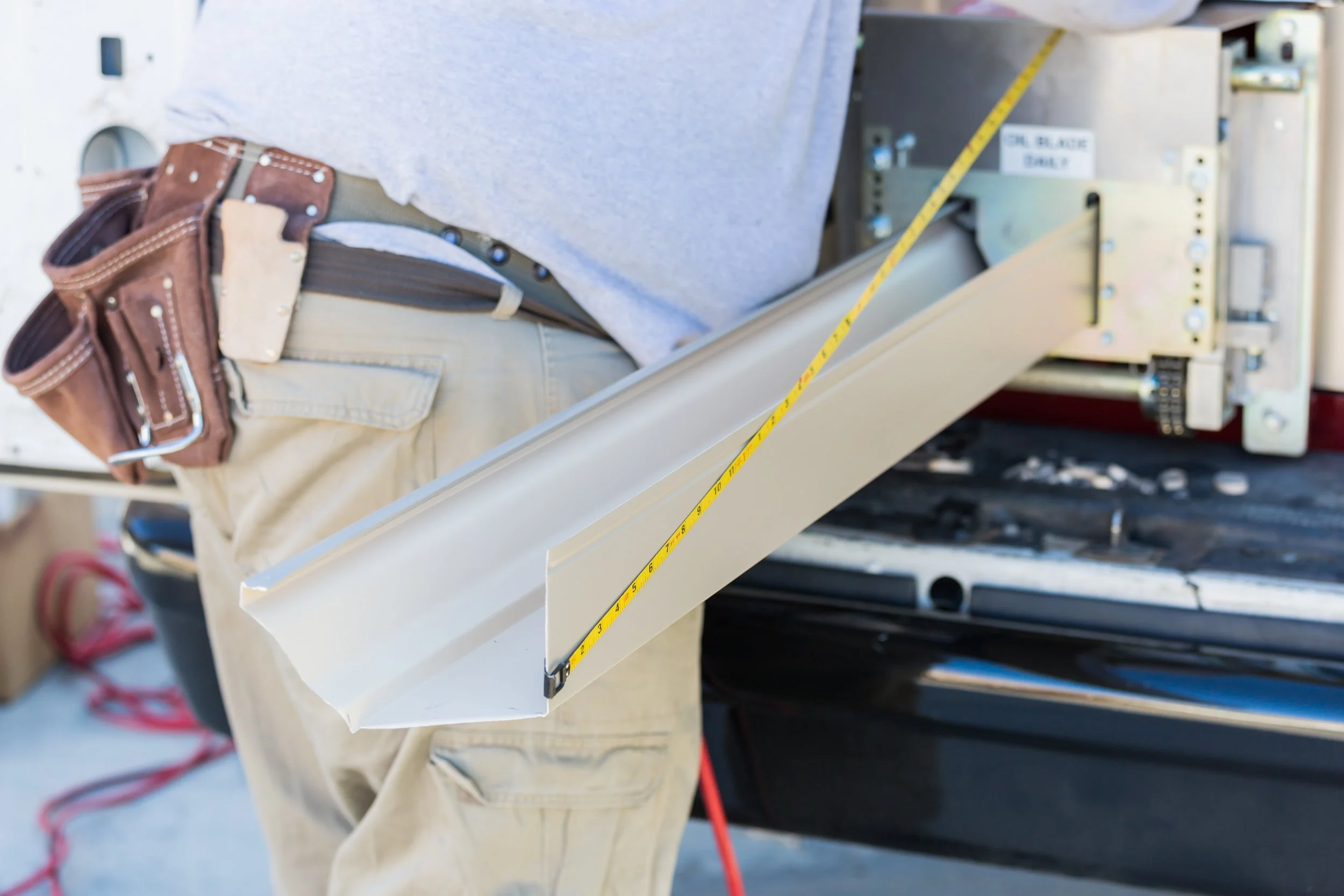 A worker in beige pants with a brown tool pouch is working on an appliance or equipment, with a metal panel and yellow measuring tape visible.