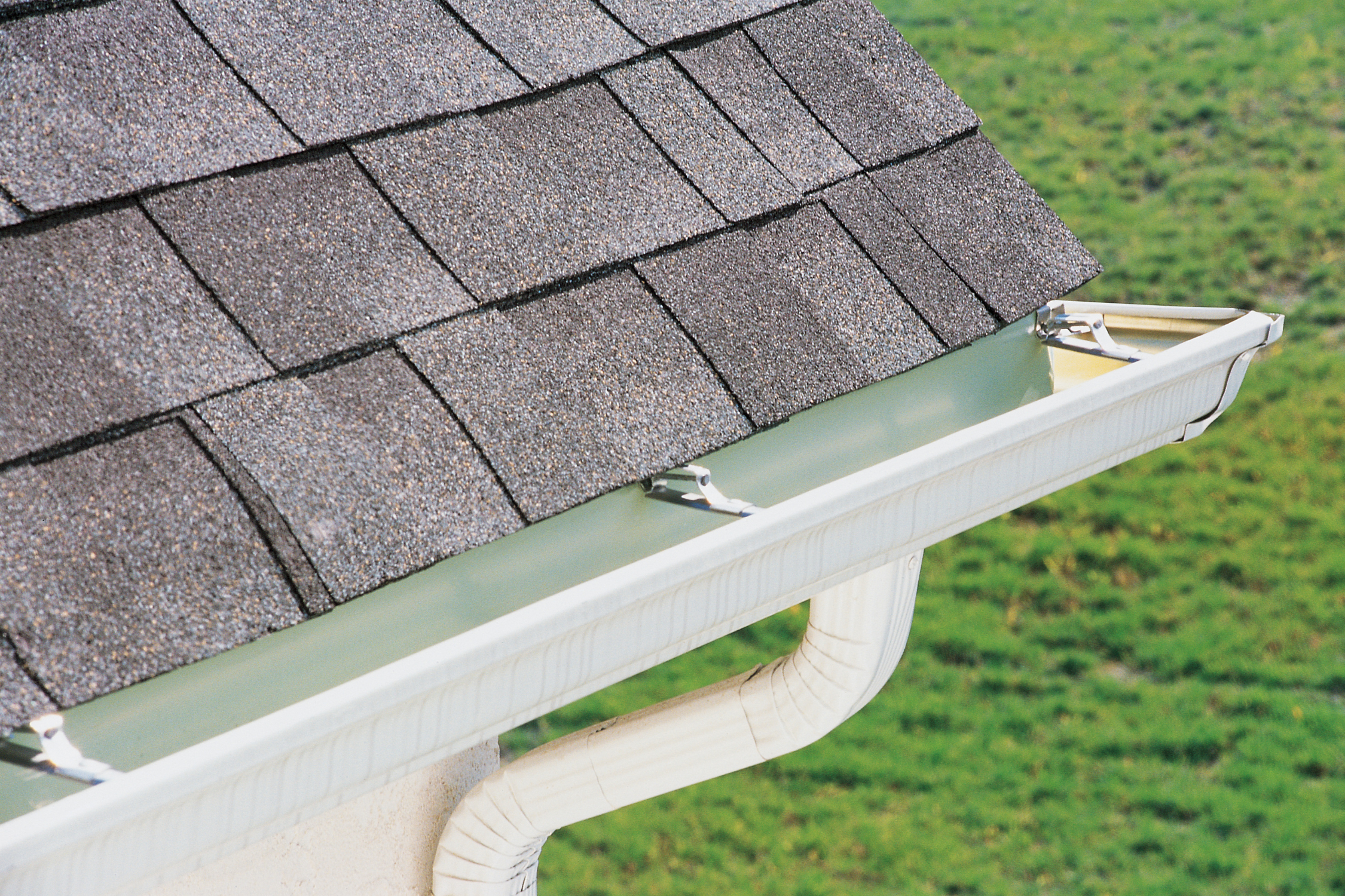 Close-up of a house roof with asphalt shingles and a white gutter system with a downspout over a grassy yard.