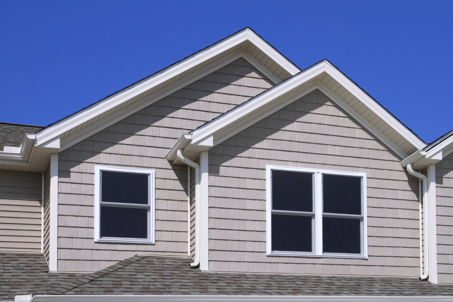 Close-up of a house's light gray siding, two double-pane windows, and a gable roof with gray shingles under a clear blue sky.