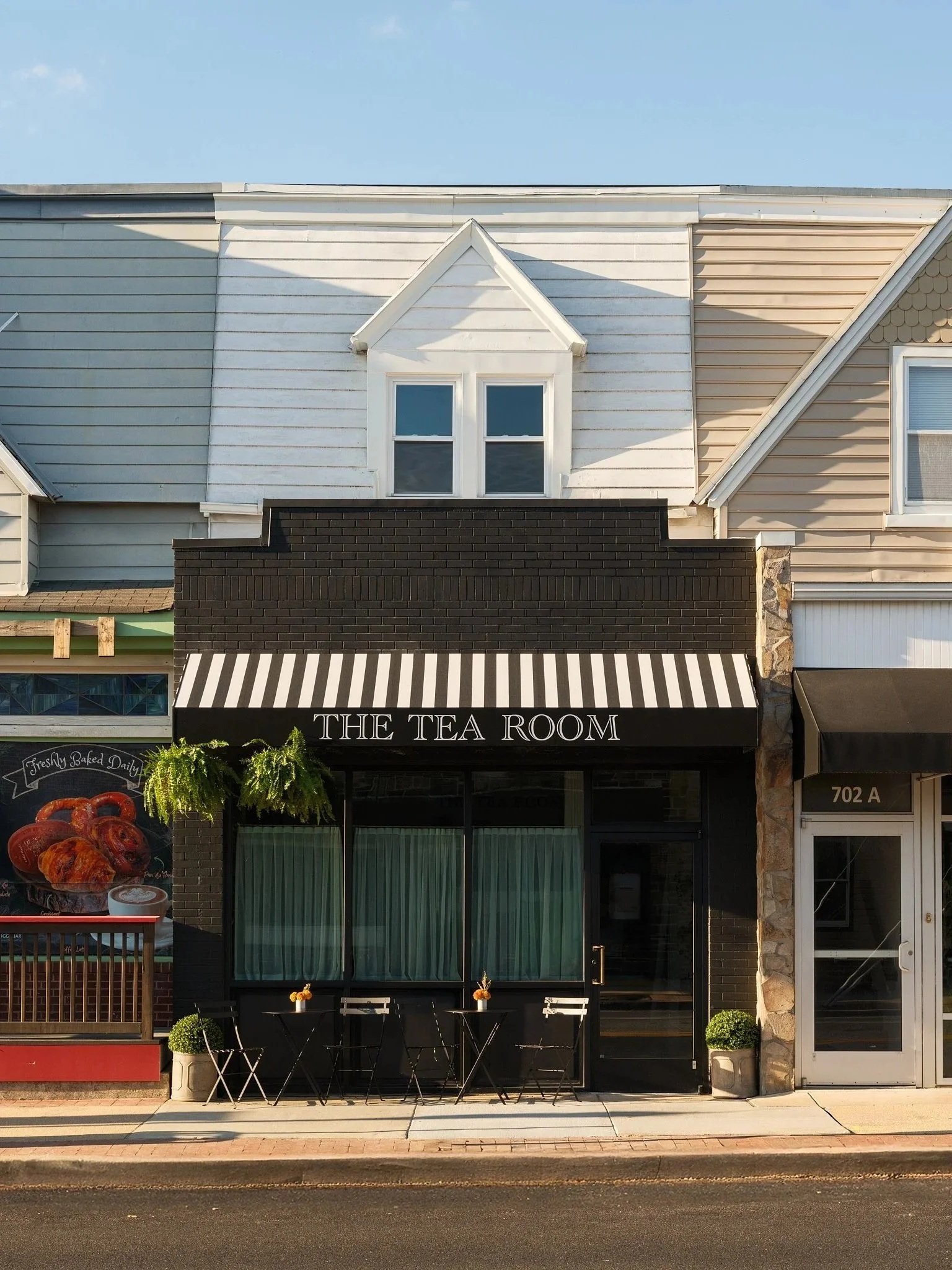  storefront with black and white striped awning labeled 'THE TEA ROOM', patio seating with three black chairs, decorative plants, and a glass door. The building has white, tan, and black exterior walls.