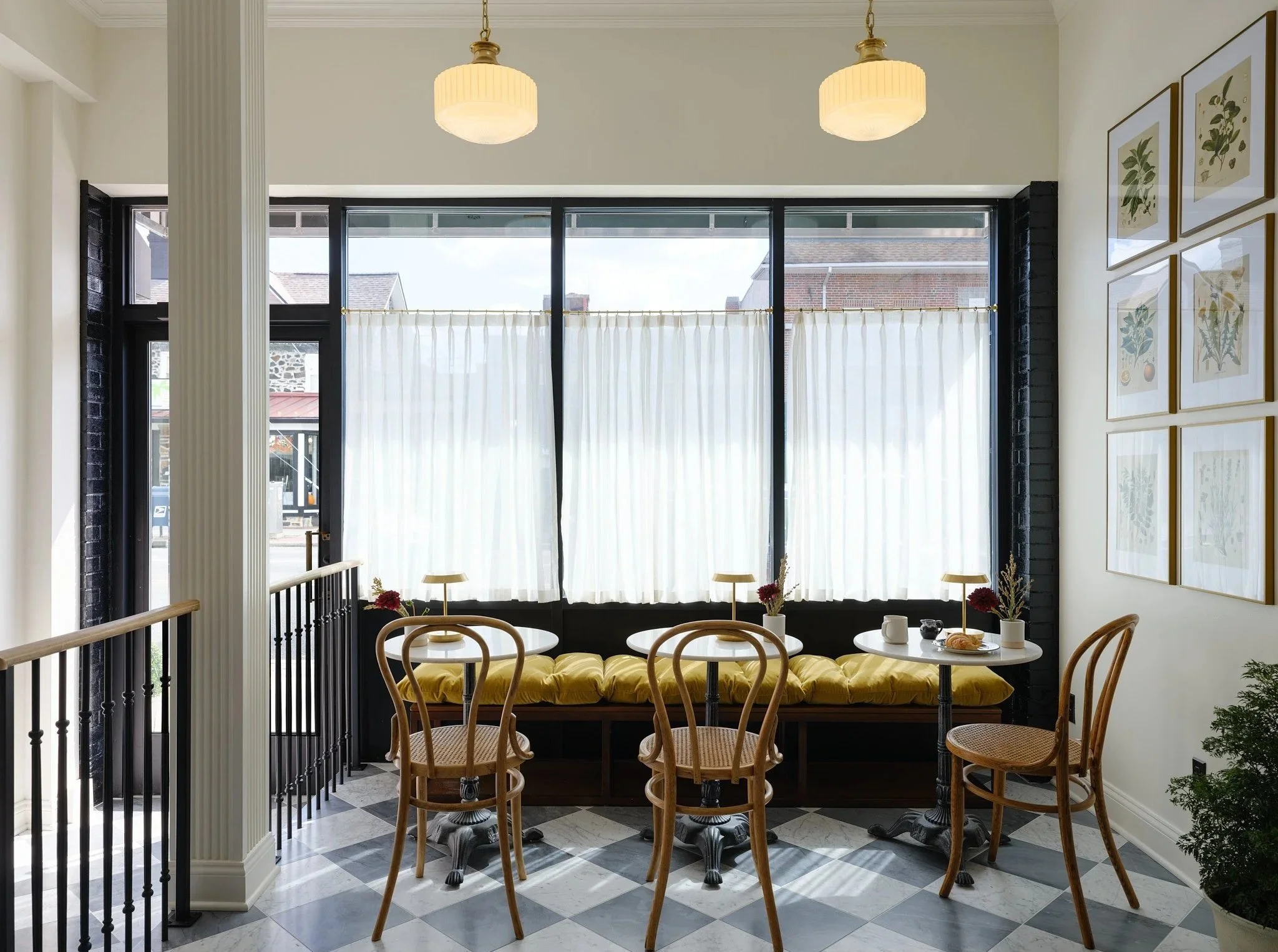 Interior of a cozy cafe with a window seat and small round tables, wooden chairs, and framed botanical prints on the wall.