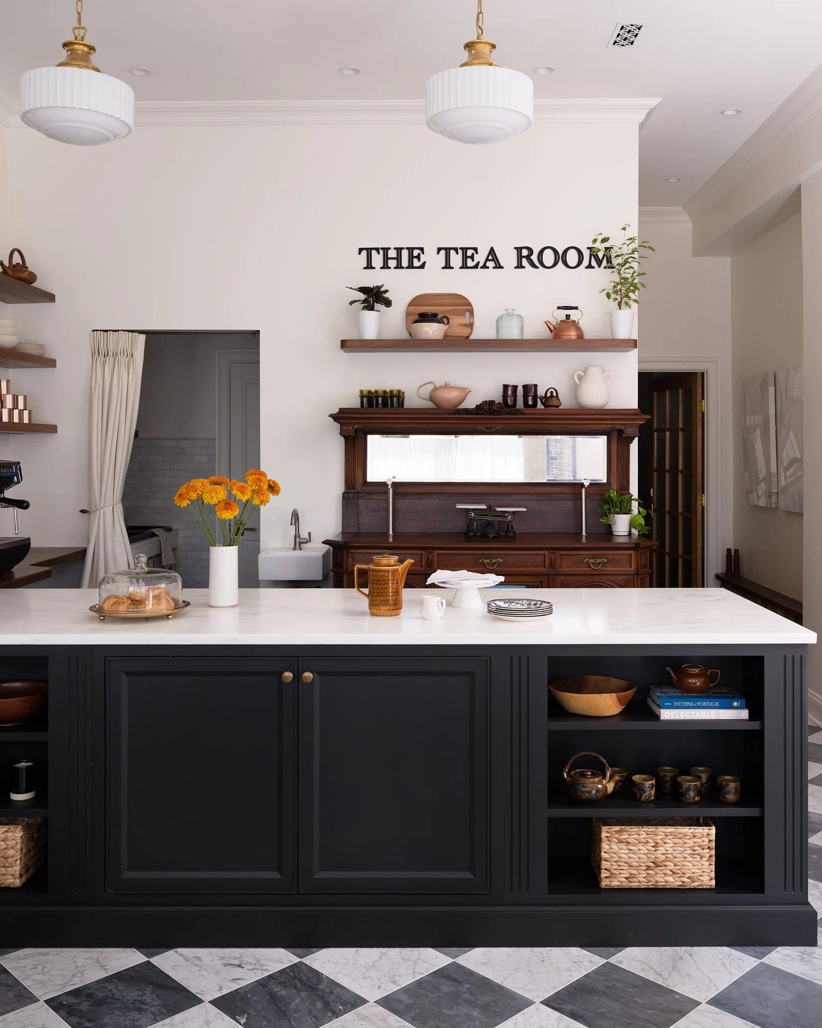 A kitchen with a white countertop, dark cabinetry, and open shelves displaying decorative items, with a sign on the wall that says 'THE TEA ROOM,' and patterned black and white tiled floor.