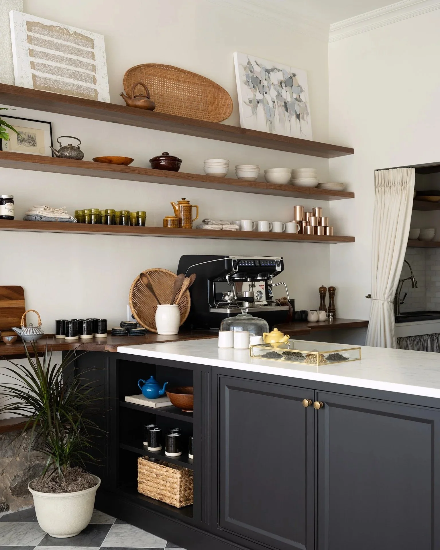 Modern kitchen with dark cabinet, open shelves displaying dishes and decor, coffee machine on counter, potted plant, and a white countertop with small kitchen items.