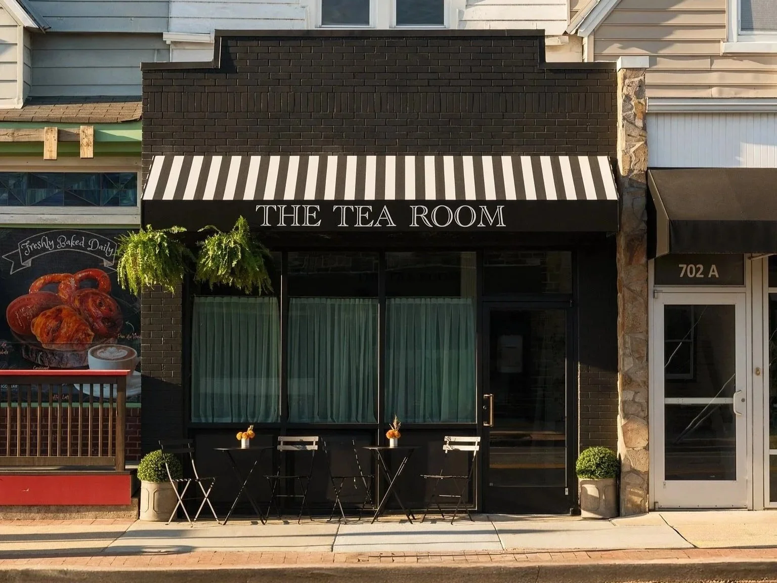 storefront with black facade, striped black and white awning labeled "The Tea Room," outdoor seating with small tables, plants, and a window with curtains.