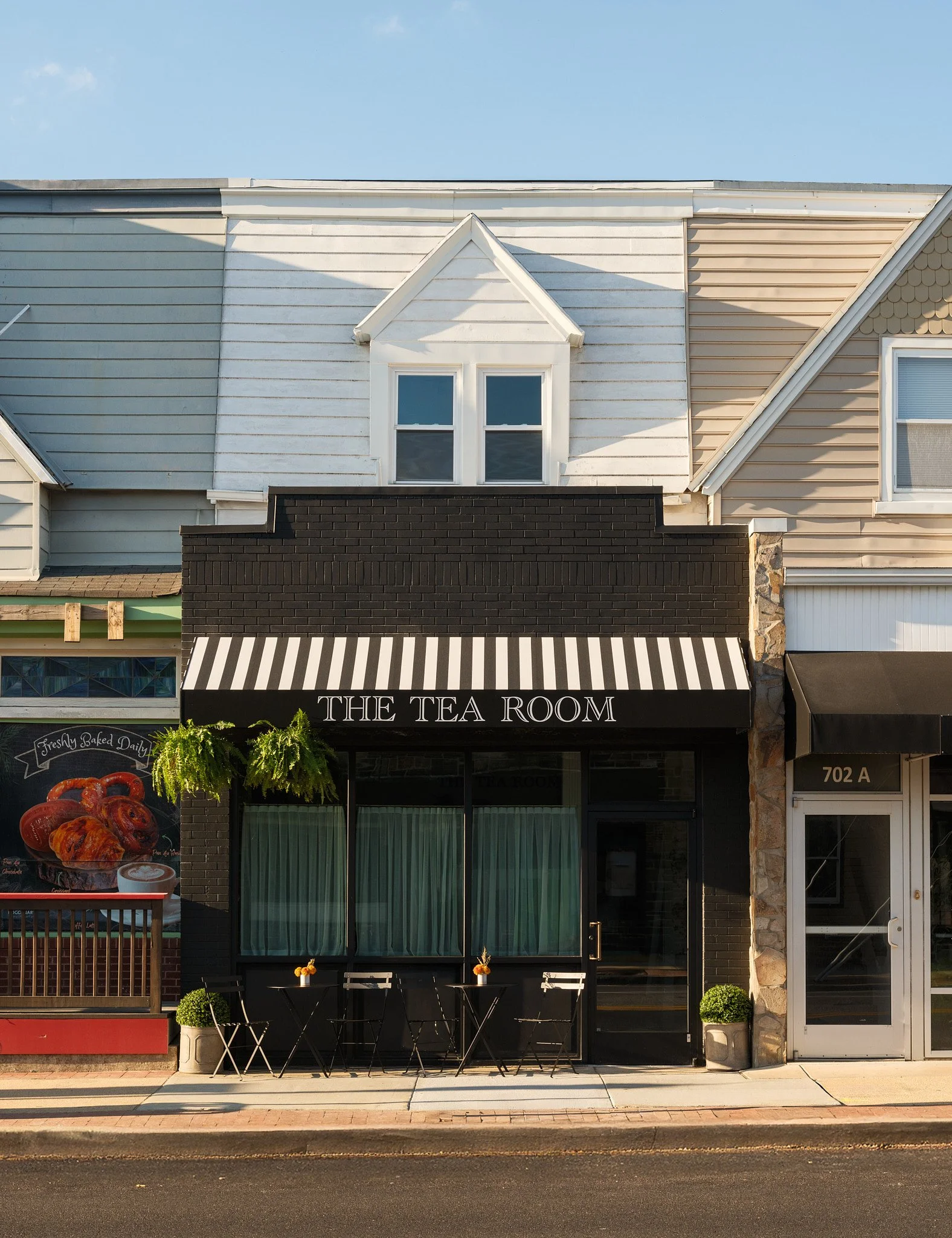 A storefront with a black awning labeled 'The Tea Room,' two tables with chairs outside, and neighboring businesses, one of which features a poster of baked goods. The building has large glass windows and doors, with small potted plants on either sid