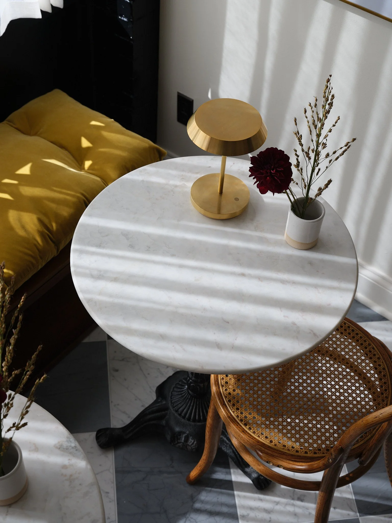 A round white marble table with a small gold lamp and a white vase with dark red flower and green foliage on it. A wooden chair with a woven cane seat is beside the table. Shadows from window blinds cast stripes across the table.