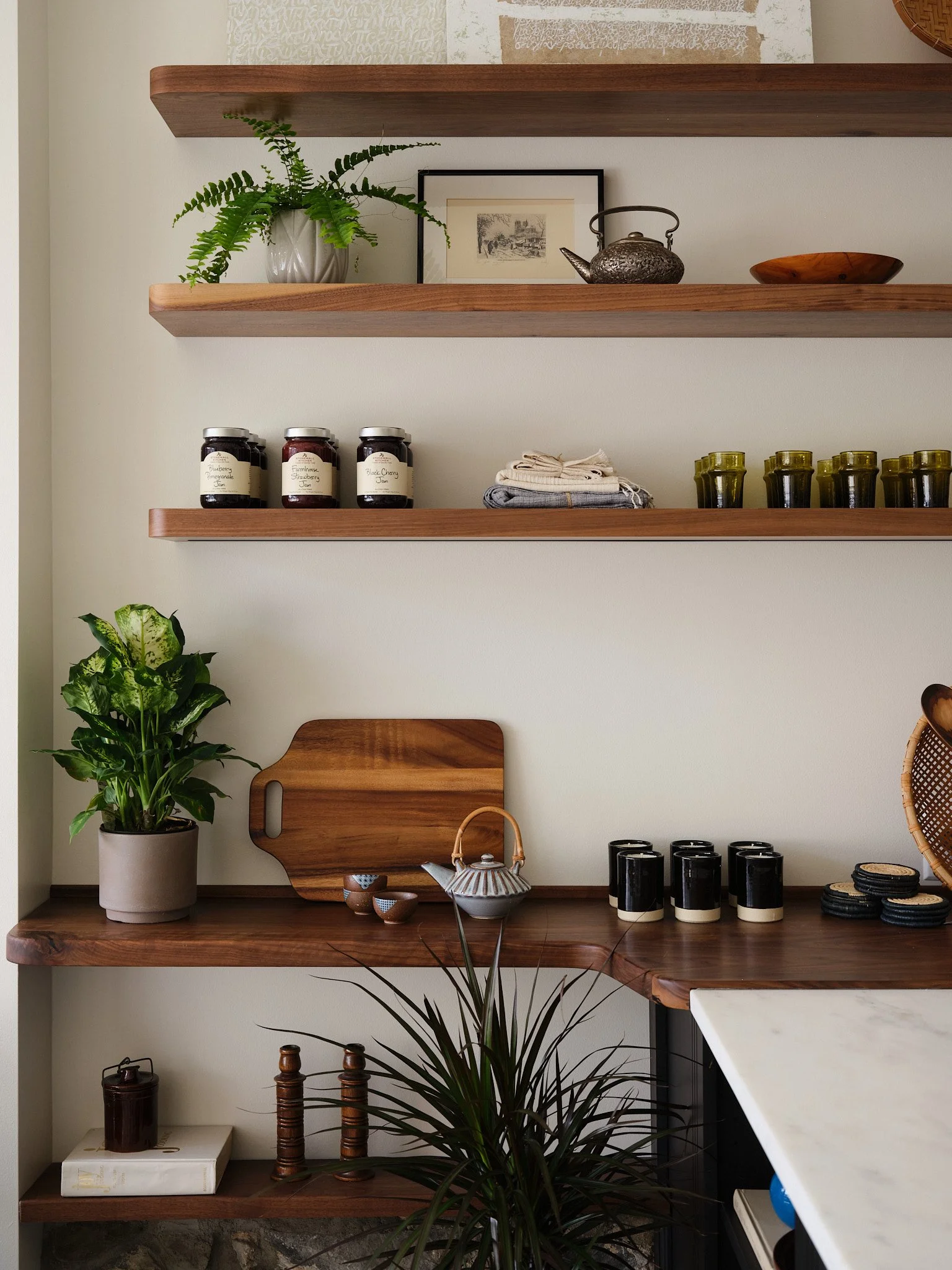 Wooden shelves on a white wall decorated with potted plants, framed artwork, teapot, dish, jars, folded cloth, cups, and ceramic dishes.