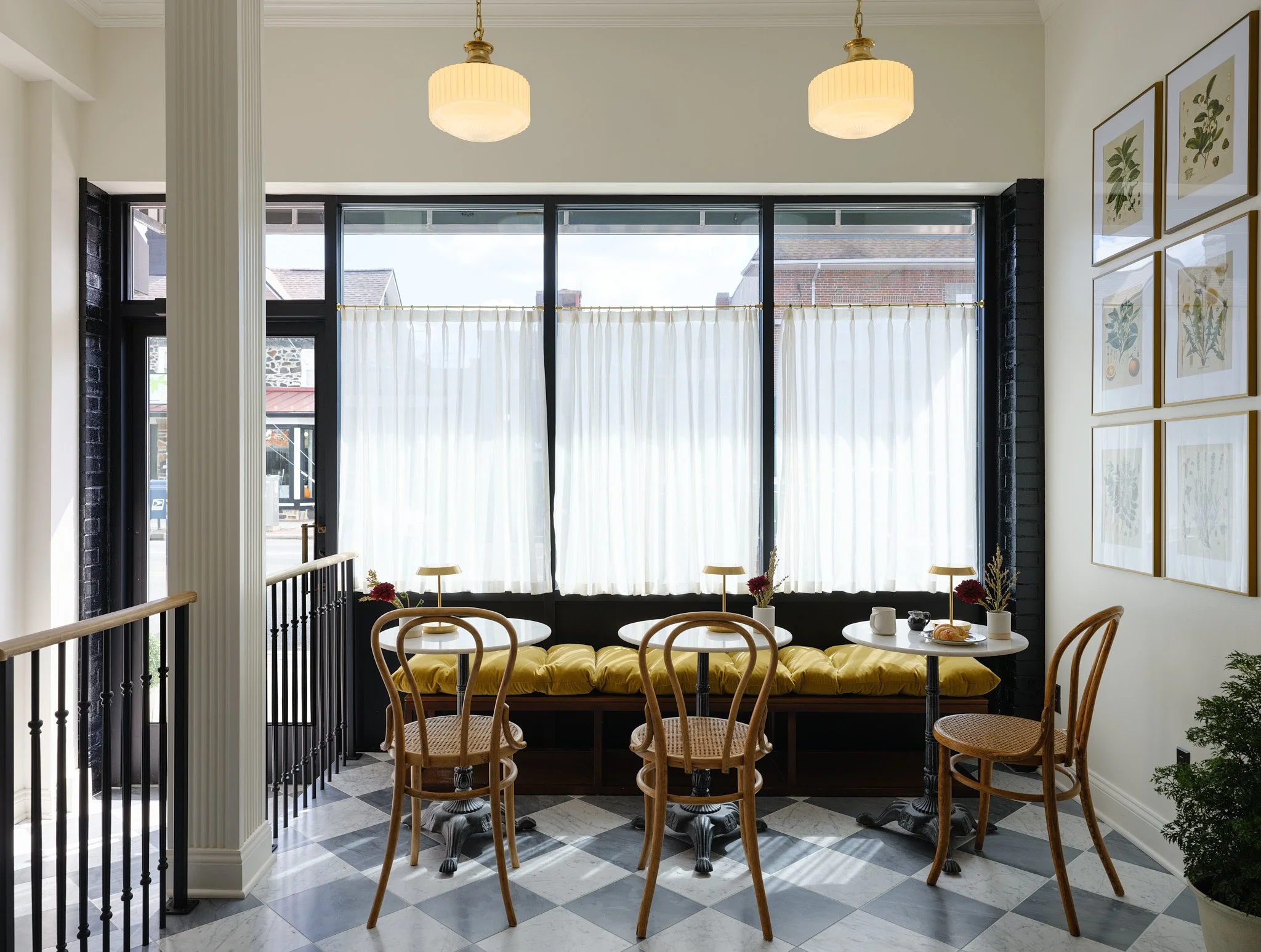 Cozy café corner with three wooden chairs, a round table, a yellow cushioned bench, and framed botanical prints on the wall, illuminated by hanging ceiling lamps and natural light from large windows with white curtains.