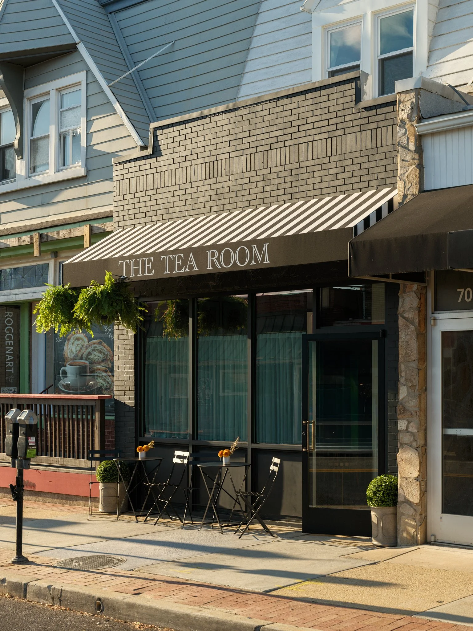 Exterior of a small cafe named 'The Tea Room' with a black and white striped awning, outdoor seating with two small tables, two white chairs, and potted plants.