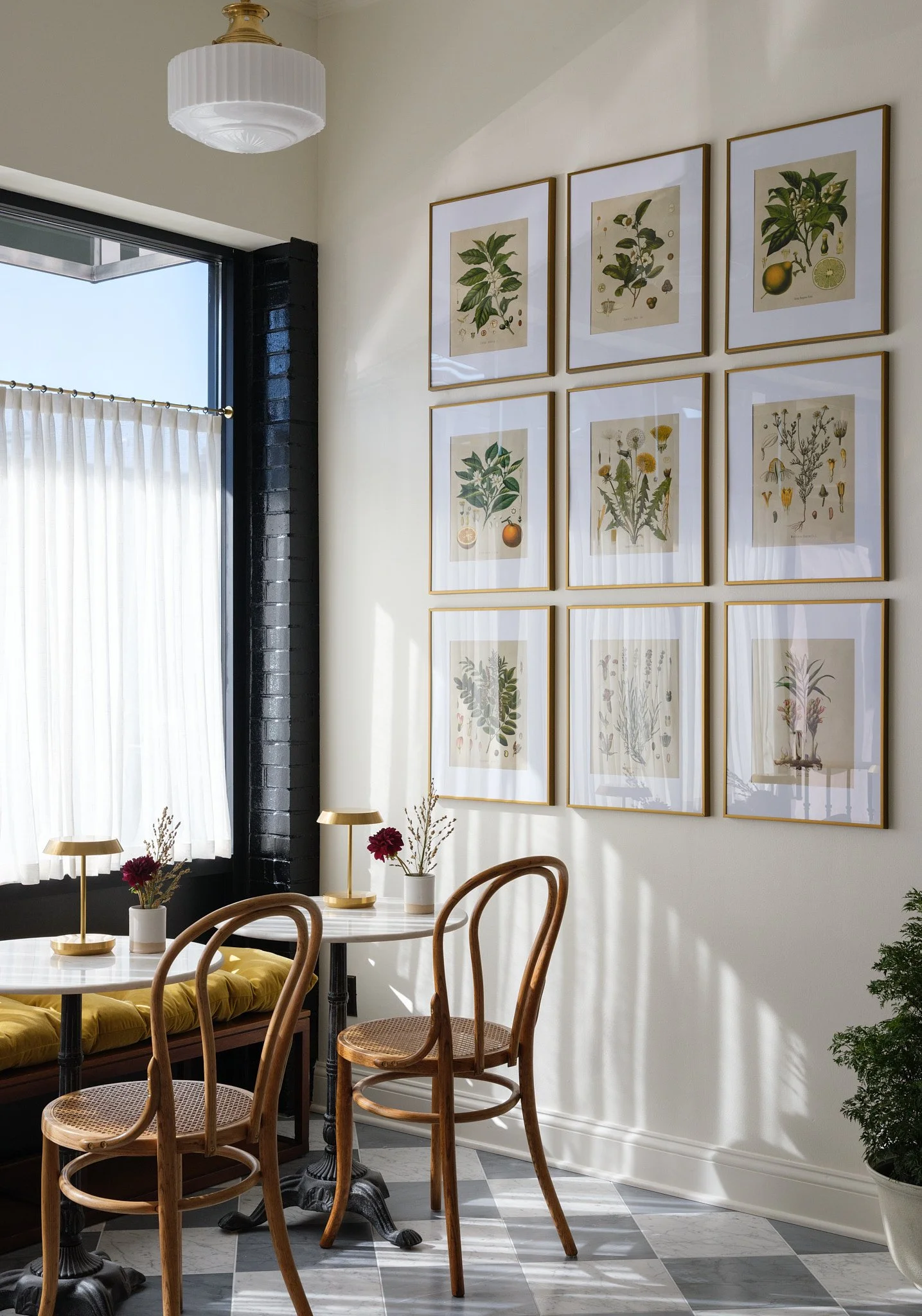 Interior of a cozy dining space with a white table, wooden chairs, framed botanical artwork on the wall, and sunlight streaming through a window with white curtains.